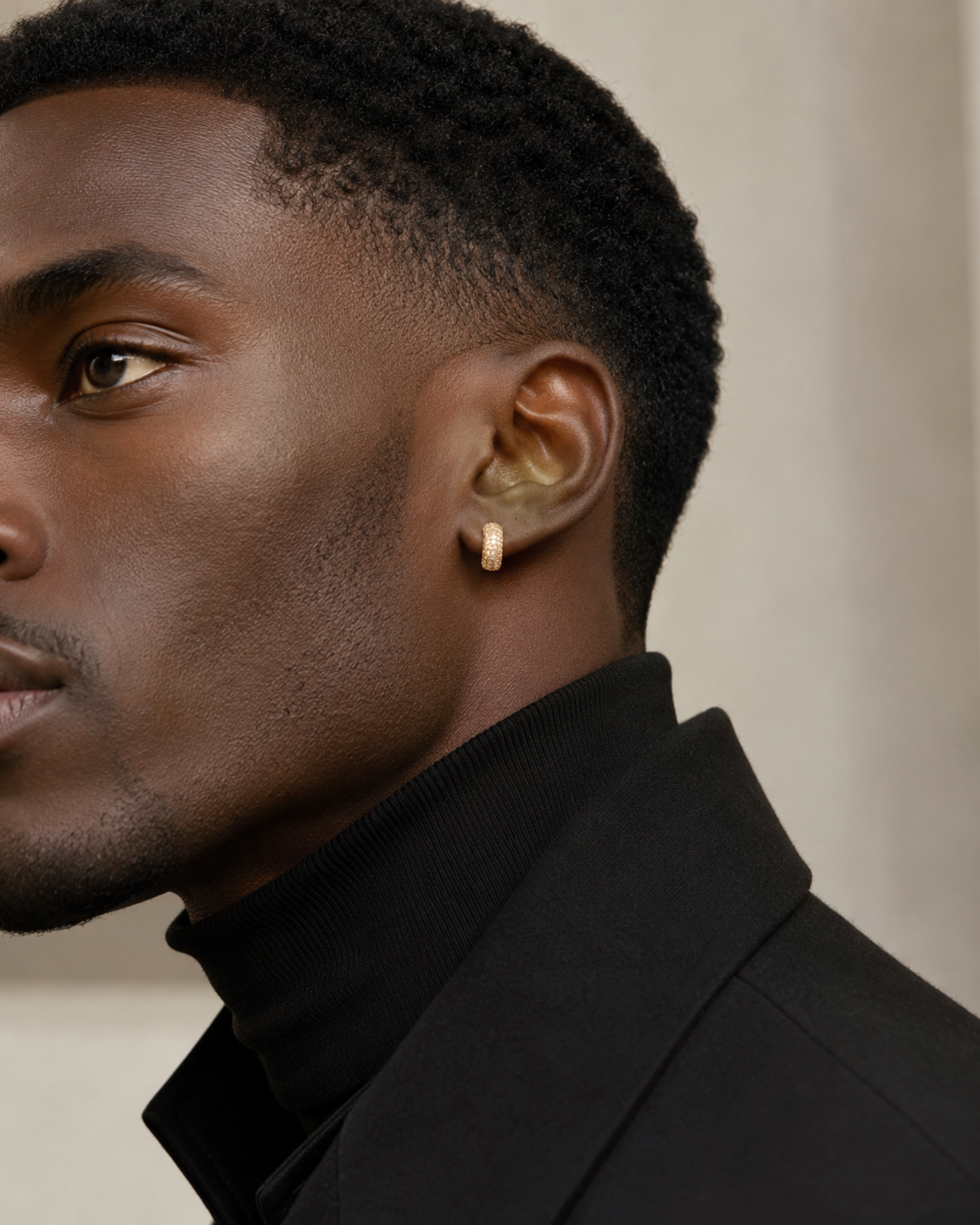 Close-up of a man's ear wearing a slim gold hoop earring, with his profile visible against a soft beige background.