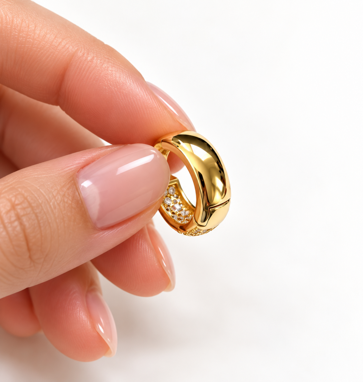 A close-up of a hand with pale pink manicured nails holding a small gold huggie hoop earring with pavé-set clear stones against a clean white background.