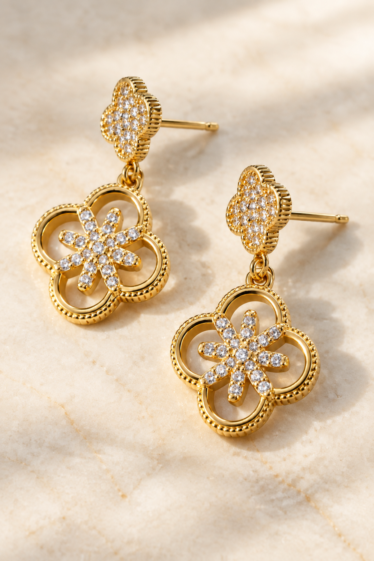 A pair of gold dangle earrings with a four-leaf clover design, adorned with pavé-set clear stones and a central flower motif, shown on a clean white background.