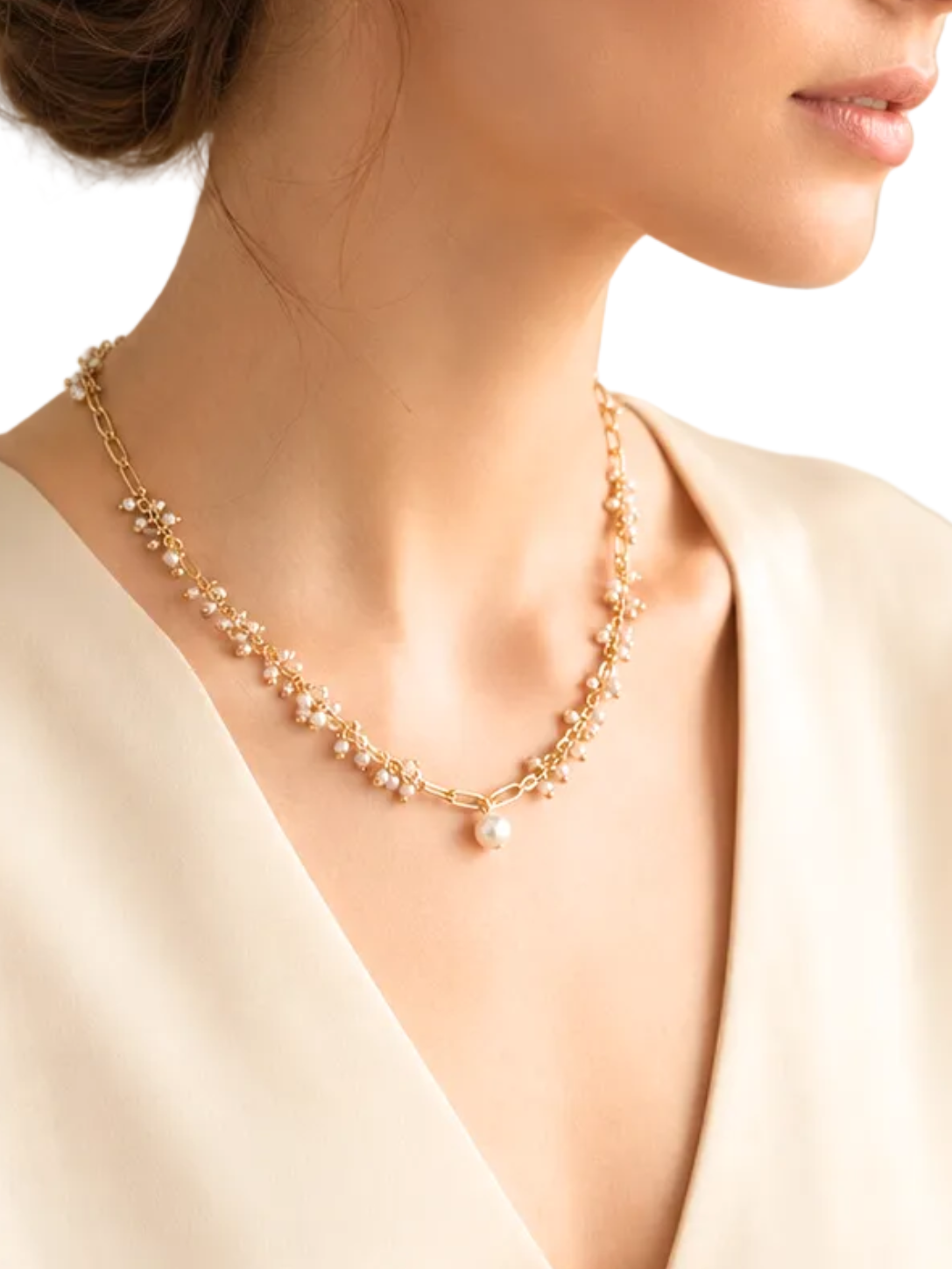 A woman in profile wearing a delicate gold beaded necklace with a small pearl pendant, styled with her hair in an elegant low bun and wearing a cream satin top against a solid white background.