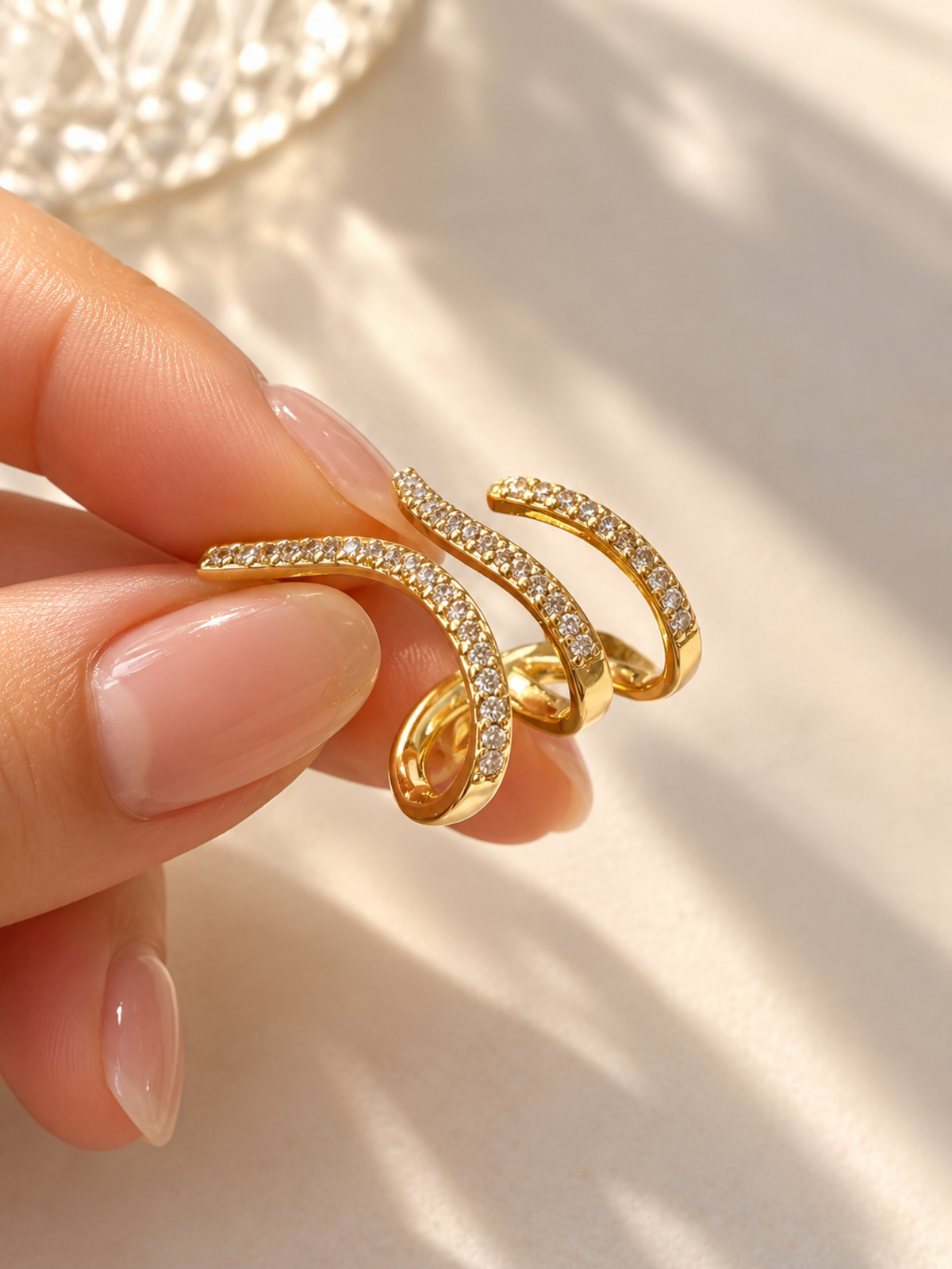 A close-up of a hand holding a gold spiral ring adorned with small clear stones, set against a warm, softly lit cream background.