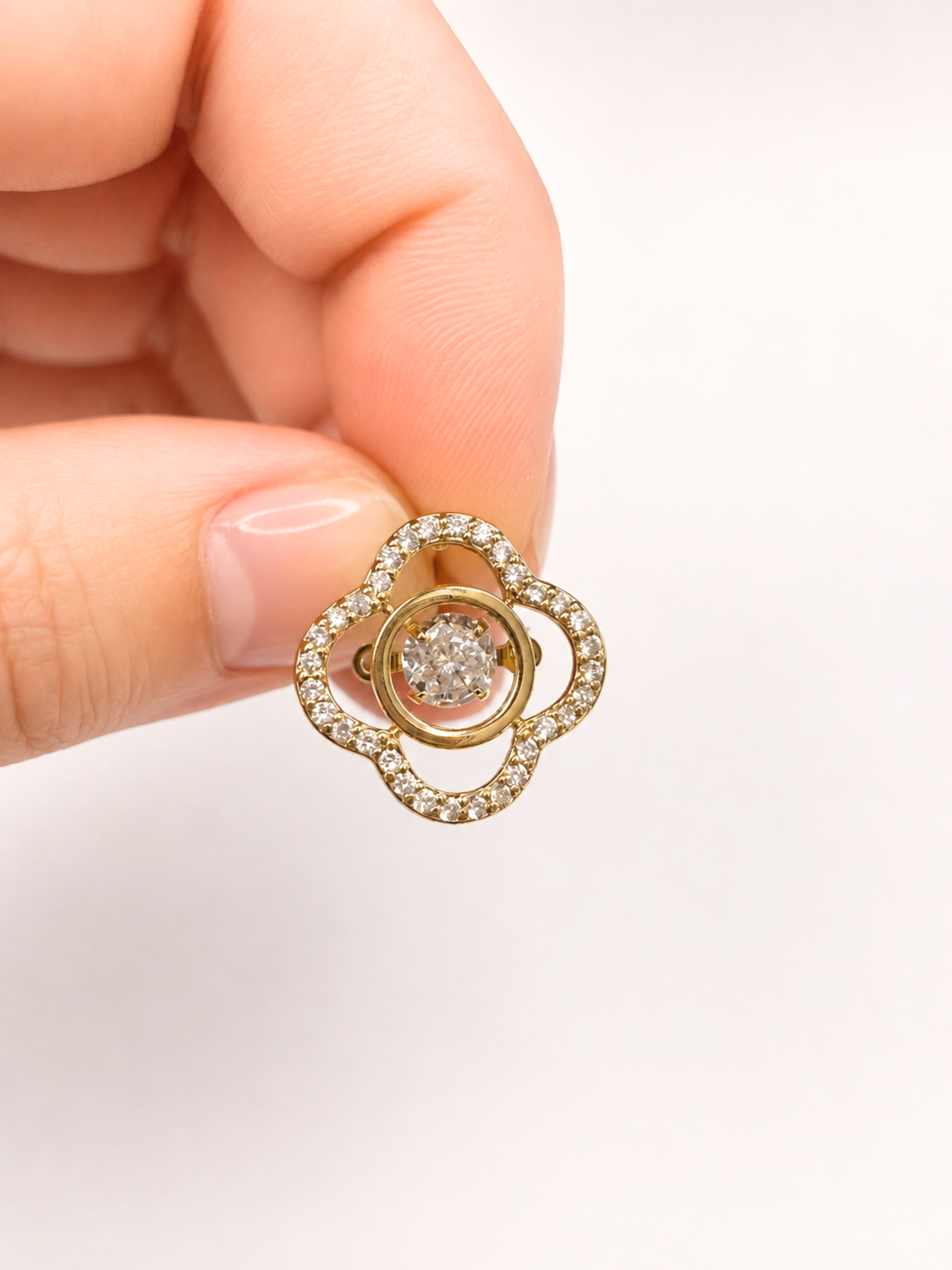 A close-up of a hand holding a small gold-toned flower-shaped ring with a central round clear gemstone and small clear stones around the outer petals, against a plain light background.
