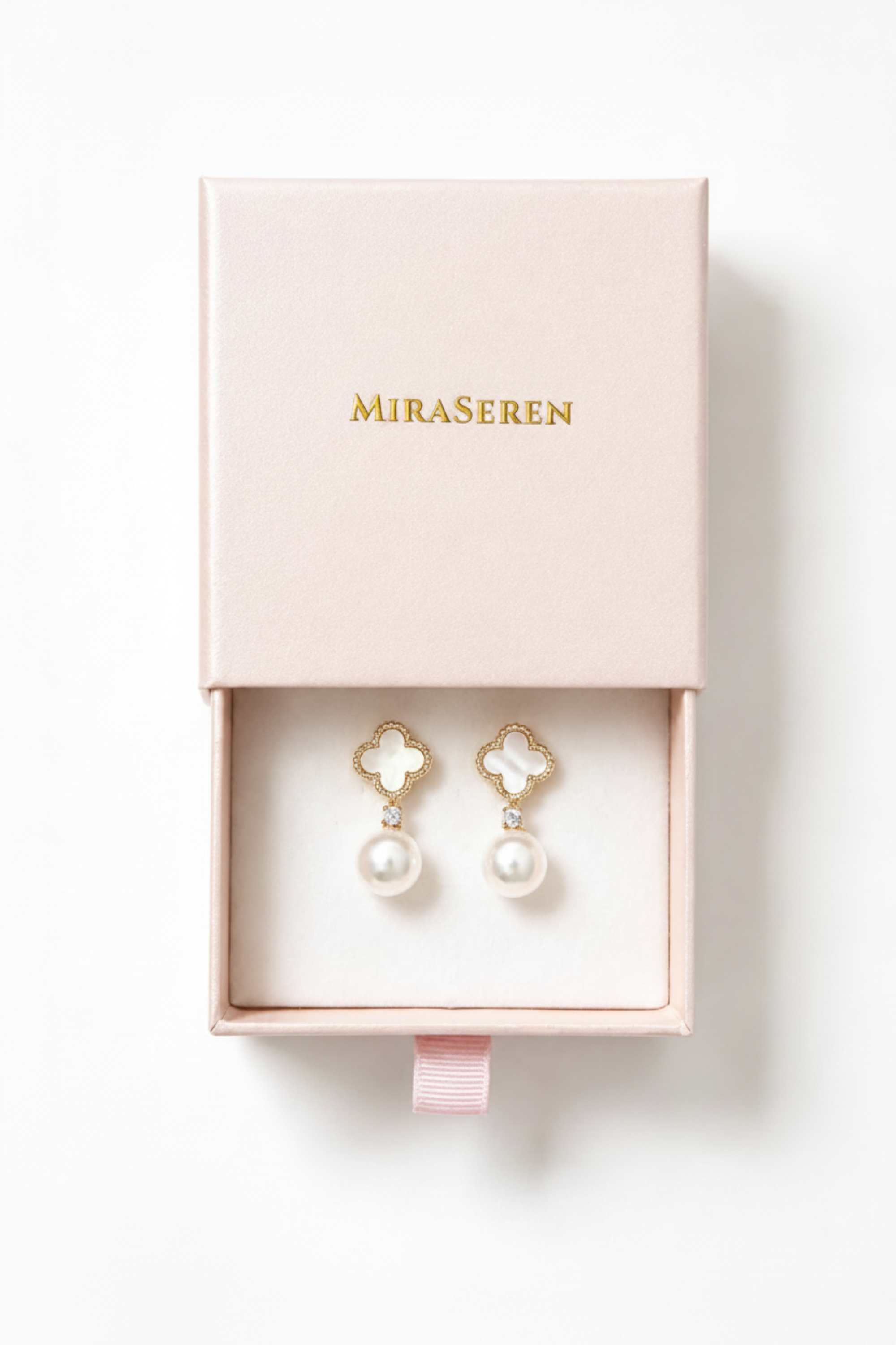 A pair of Miraseren drop earrings with white mother-of-pearl pearls and gold clover-shaped settings, presented in an open pale pink jewelry box on a white background.