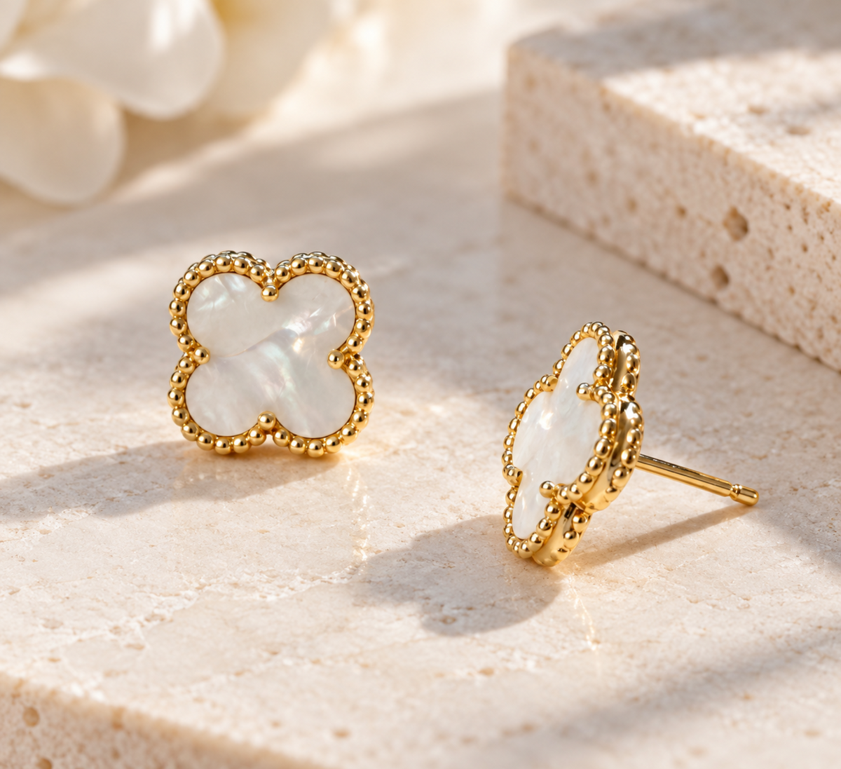 A pair of gold-toned four-leaf clover stud earrings with iridescent white mother-of-pearl inlays, displayed on a textured beige stone surface with soft shadows.
