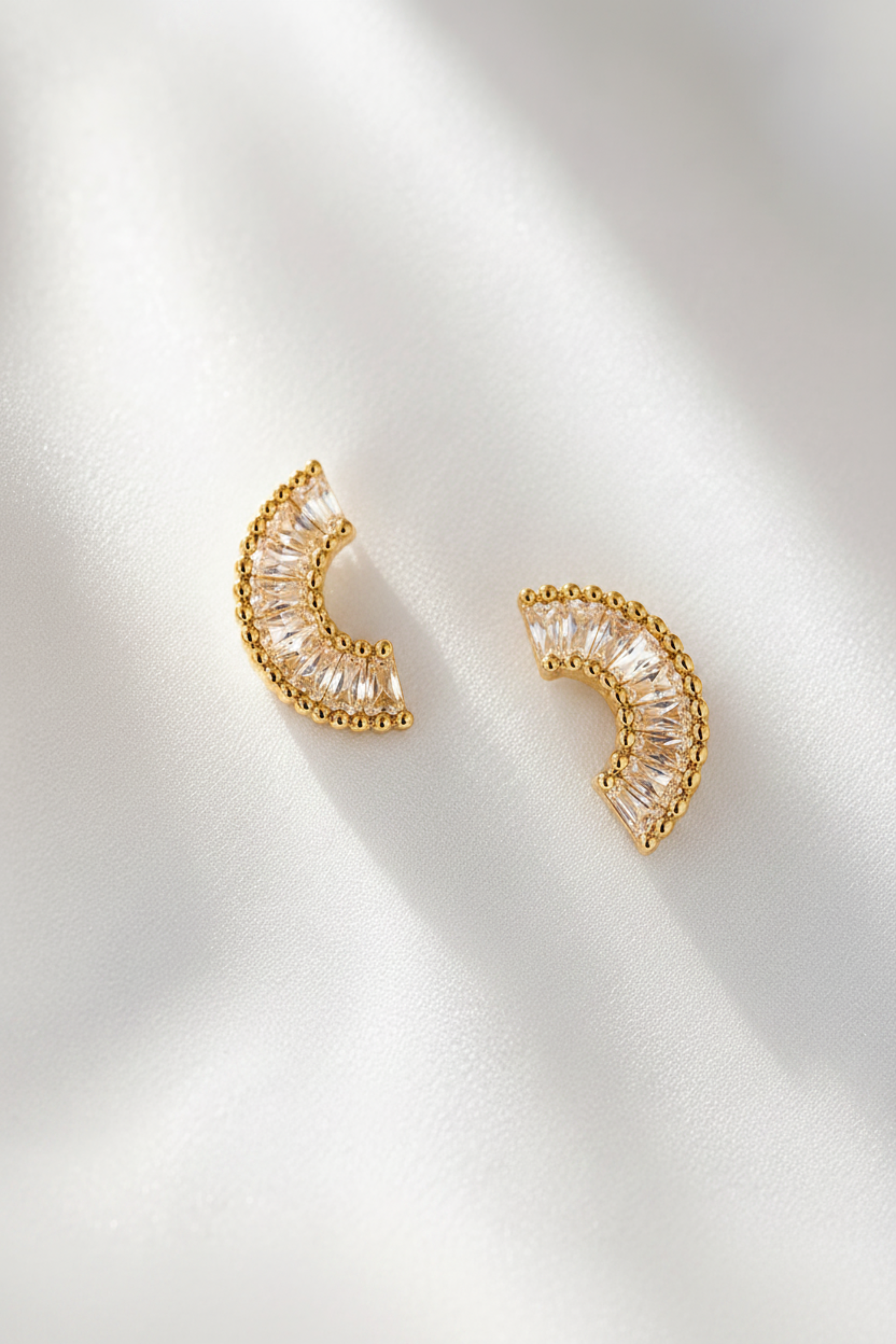A pair of gold crescent-shaped stud earrings with clear baguette-cut stones, displayed on a warm beige satin fabric background.
