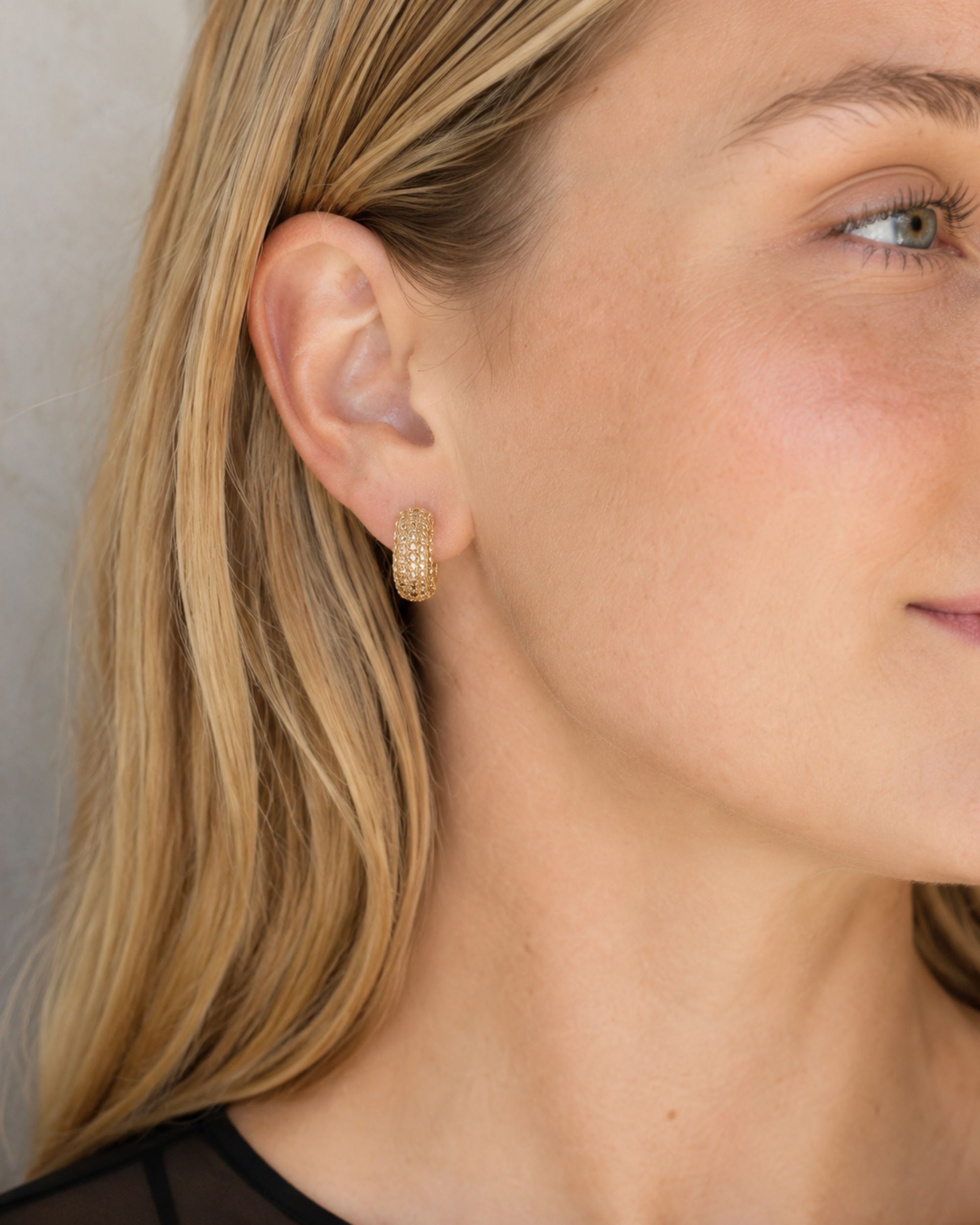 A close-up side profile of a woman with blonde hair wearing a small gold hoop earring with a textured, pave-like surface.