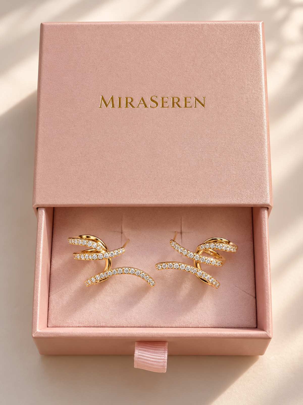 A pair of gold wrap-style earrings with rows of clear pavé stones, displayed inside an open pink Miraseren jewelry box on a soft pink velvet insert.