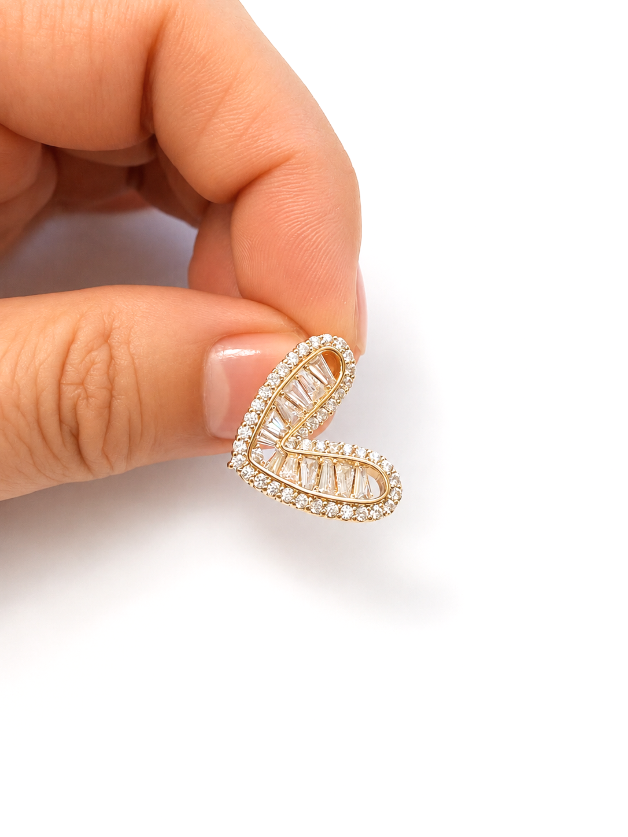 A close-up of a hand holding a small gold heart-shaped stud earring paved with clear stones against a clean white background.
