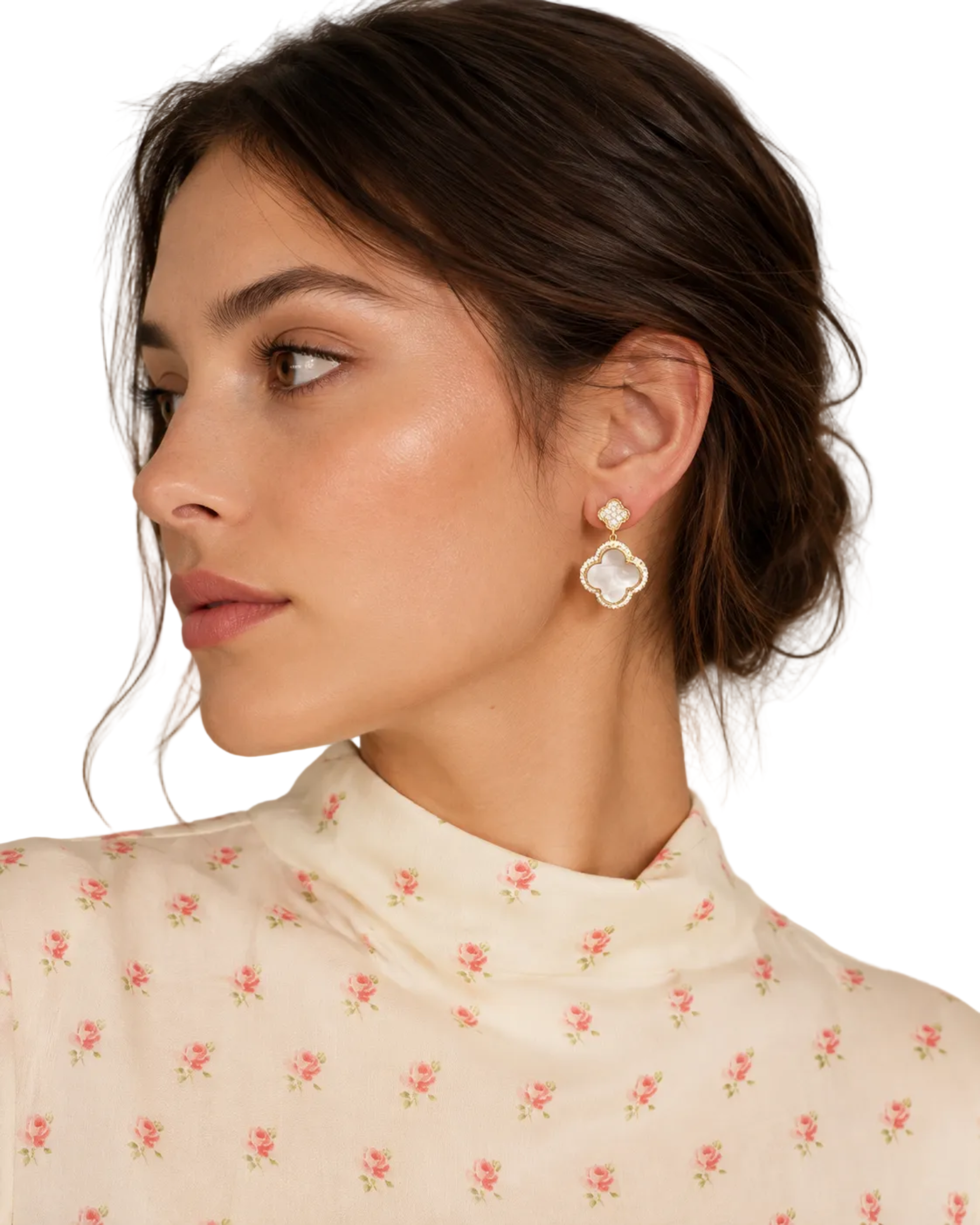 A close-up side profile of a woman with dark hair in a low bun, wearing a cream floral mock neck top and a gold four-petal drop earring with a white pearl center.