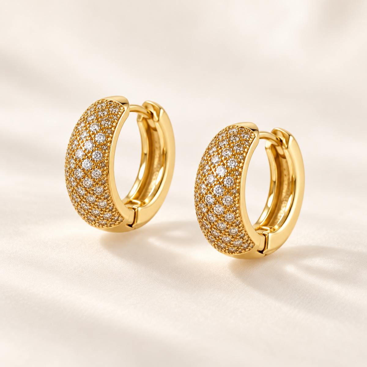 A pair of gold huggie hoop earrings paved with small clear stones, displayed on a soft, light beige fabric background.