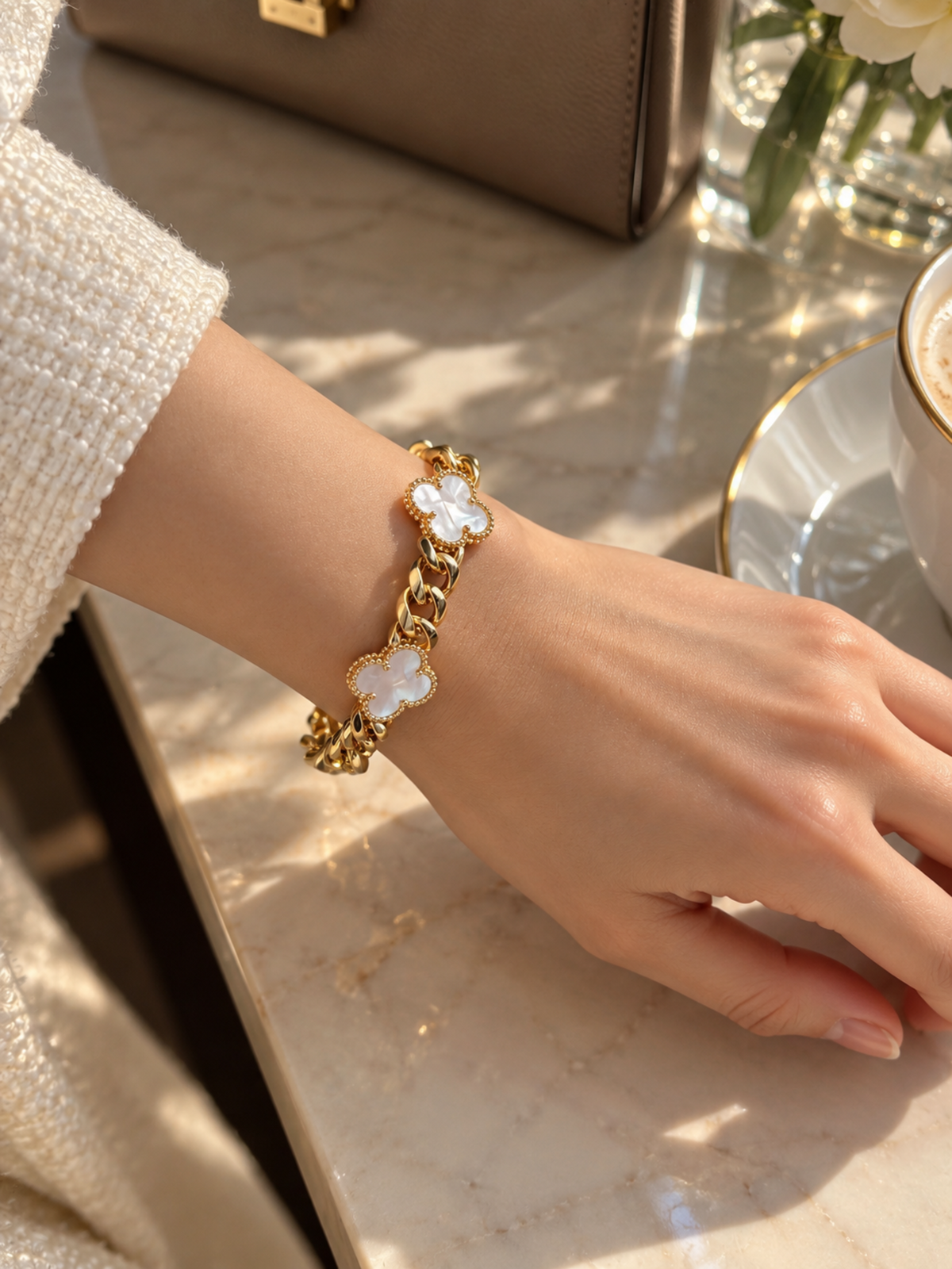 A gold-tone chain bracelet with four white mother-of-pearl clover charms, worn on a woman's wrist in a bright, sunlit indoor setting.