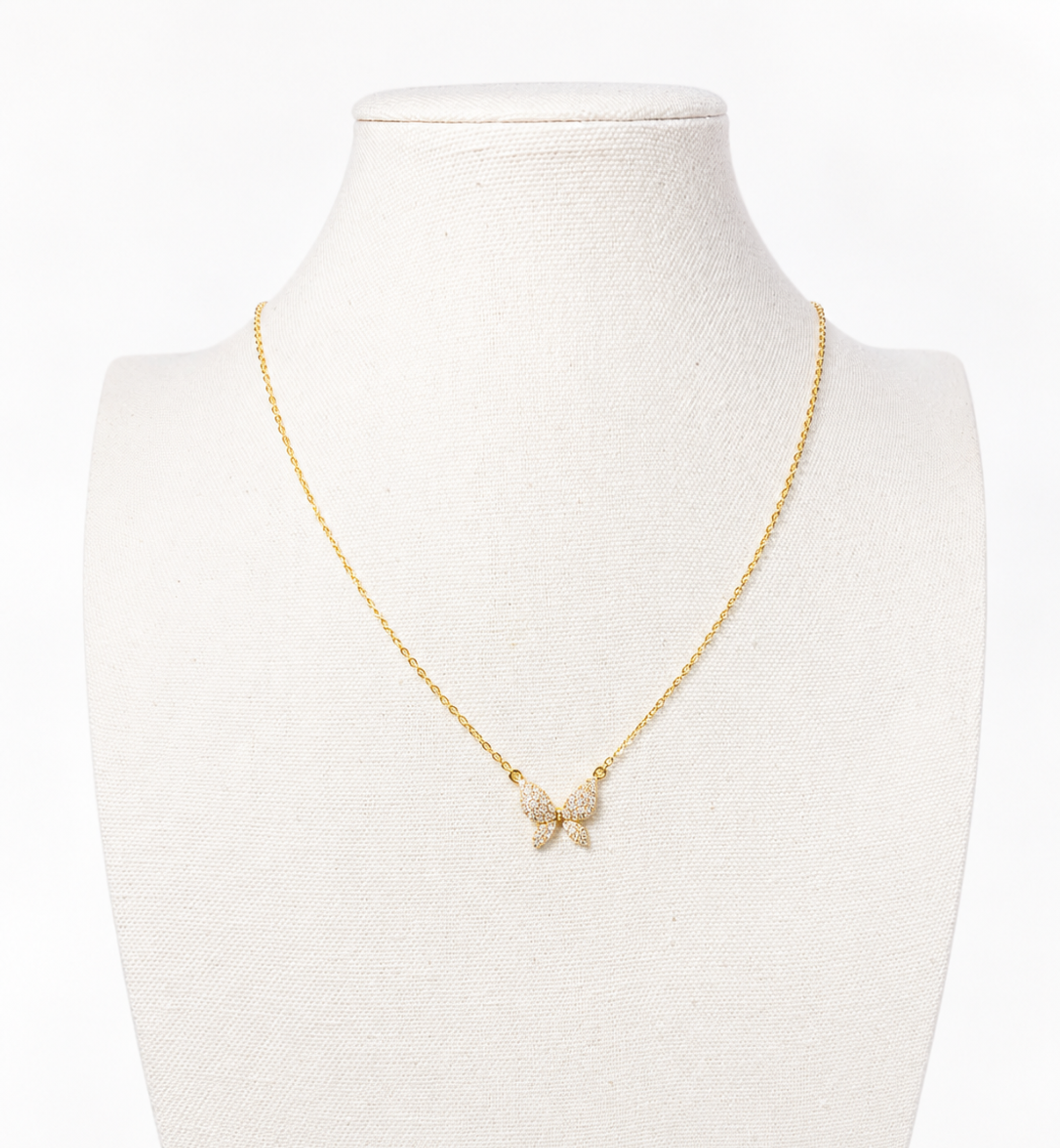 A delicate gold chain necklace with a small bow-shaped pendant adorned with clear rhinestones, displayed on a cream-colored jewelry bust against a white background.