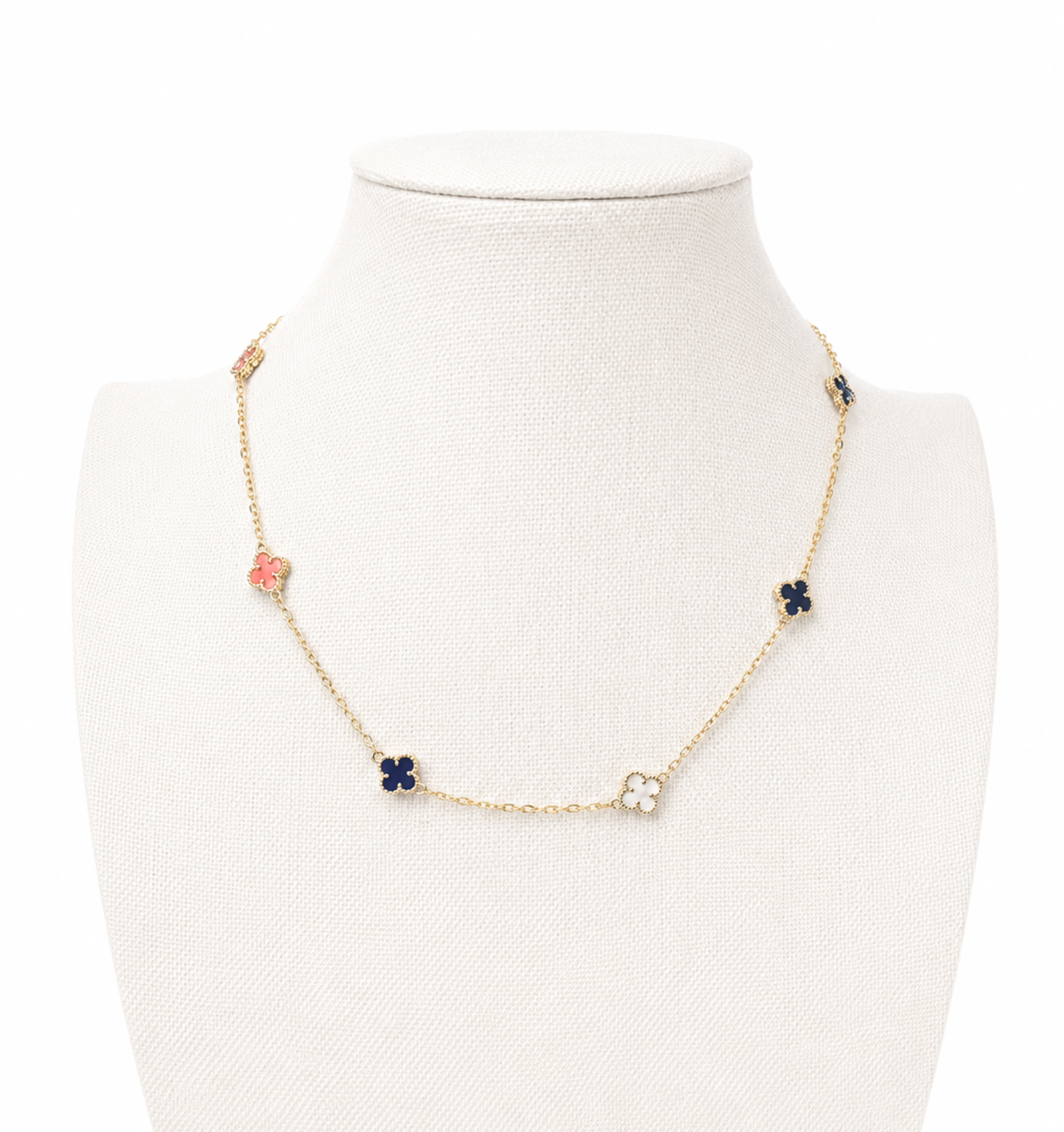 A delicate gold chain necklace displayed on a cream-colored jewelry bust. The necklace features small floral clover motifs in navy blue, pink, and white, set with tiny clear stones.