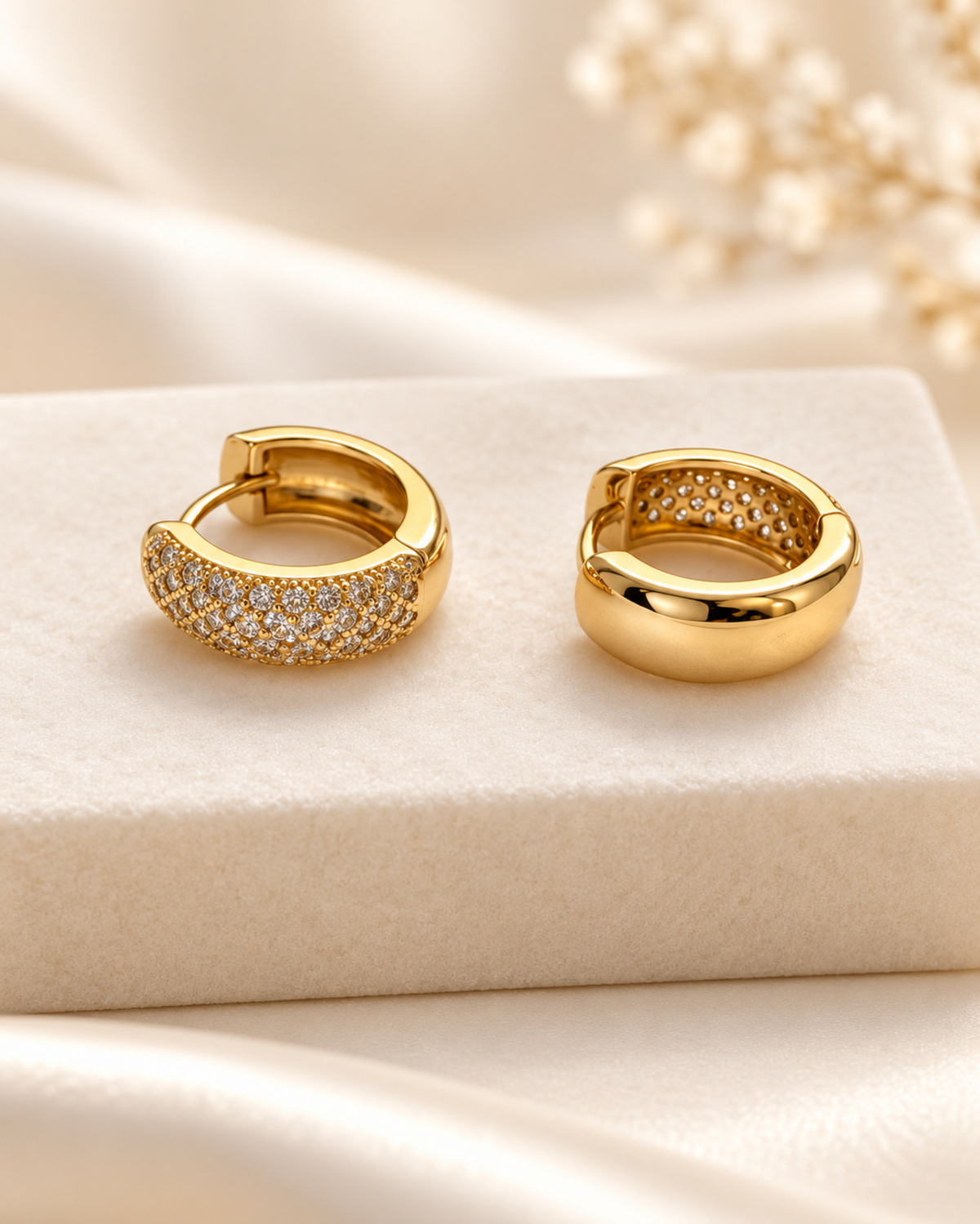 A pair of gold huggie hoop earrings displayed on a cream-colored velvet jewelry box. One earring features a pavé-set row of clear stones, while the other is a polished solid gold.