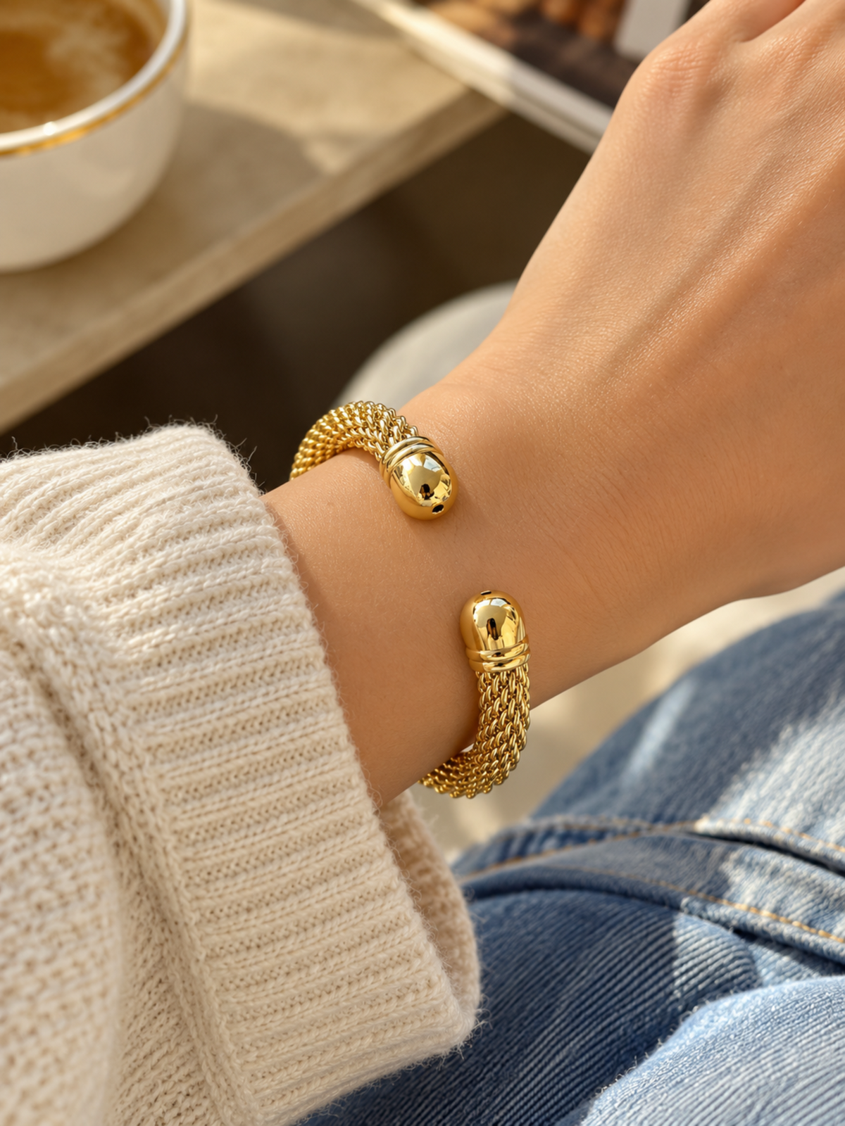 A close-up of a person's wrist wearing a gold snake-head cuff bracelet, styled with a cream knit sweater and blue jeans in a warm indoor setting.
