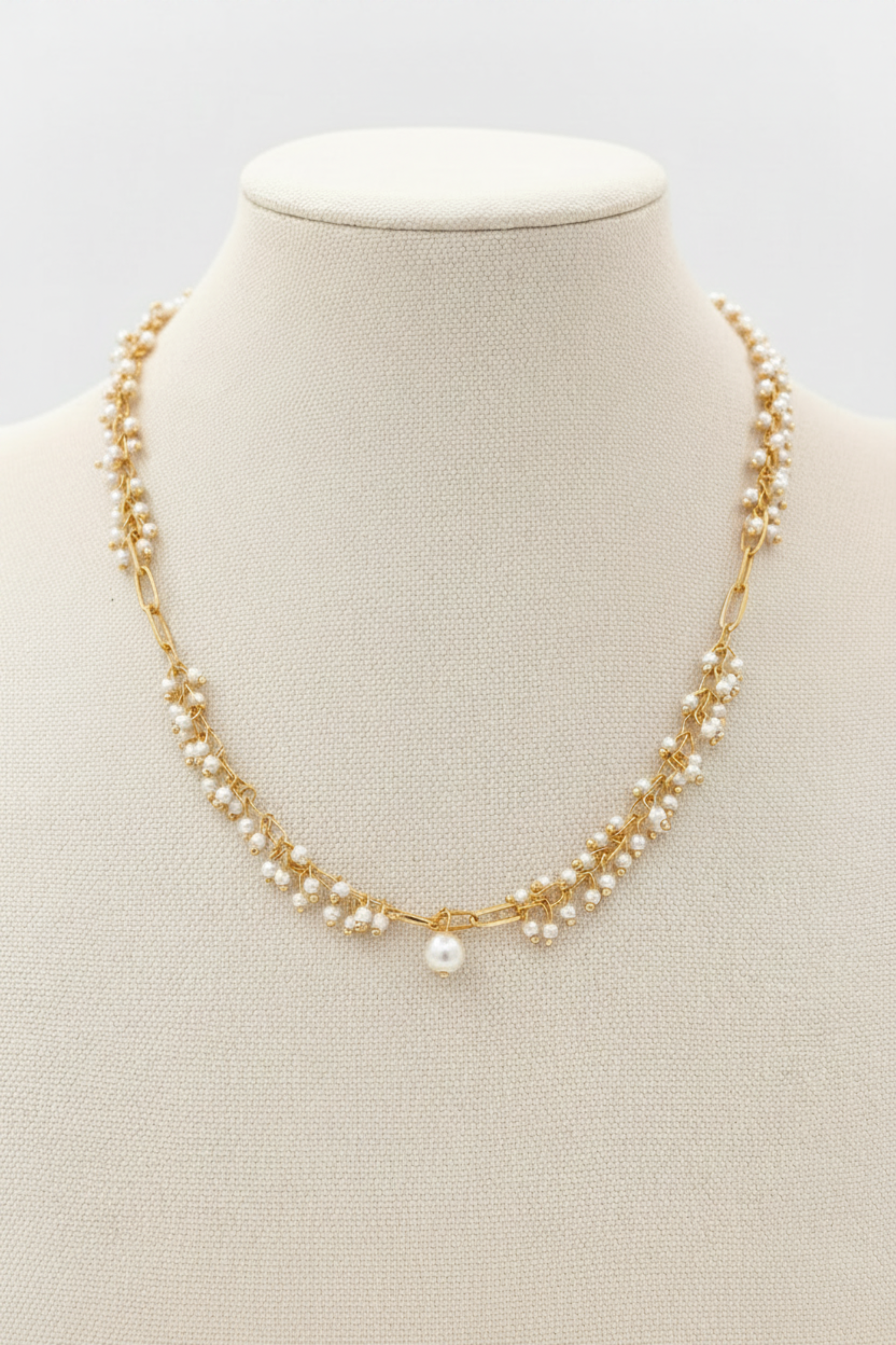 A gold chain necklace with multiple strands of small white pearls and a single larger pearl pendant, displayed on a cream fabric jewelry bust.