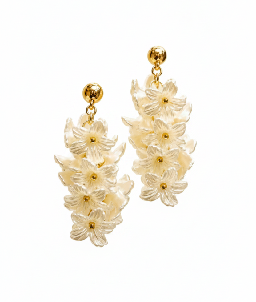 A pair of dangling floral earrings with layered translucent white flower clusters and polished gold-tone posts and centers, shown against a soft warm white bokeh background.