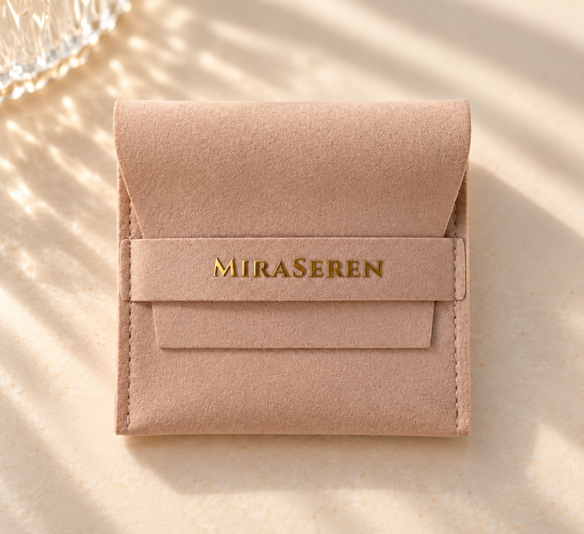 A small blush pink suede jewelry pouch with a gold-tone 'MIRA SEREN' logo on the front, photographed on a warm beige surface with soft leaf shadows.