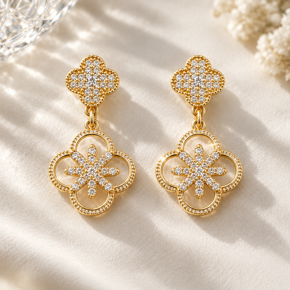 A pair of gold-toned drop earrings with four-leaf clover motifs, embellished with clear pavé stones and a central starburst design, displayed on a warm, sunlit cream surface.