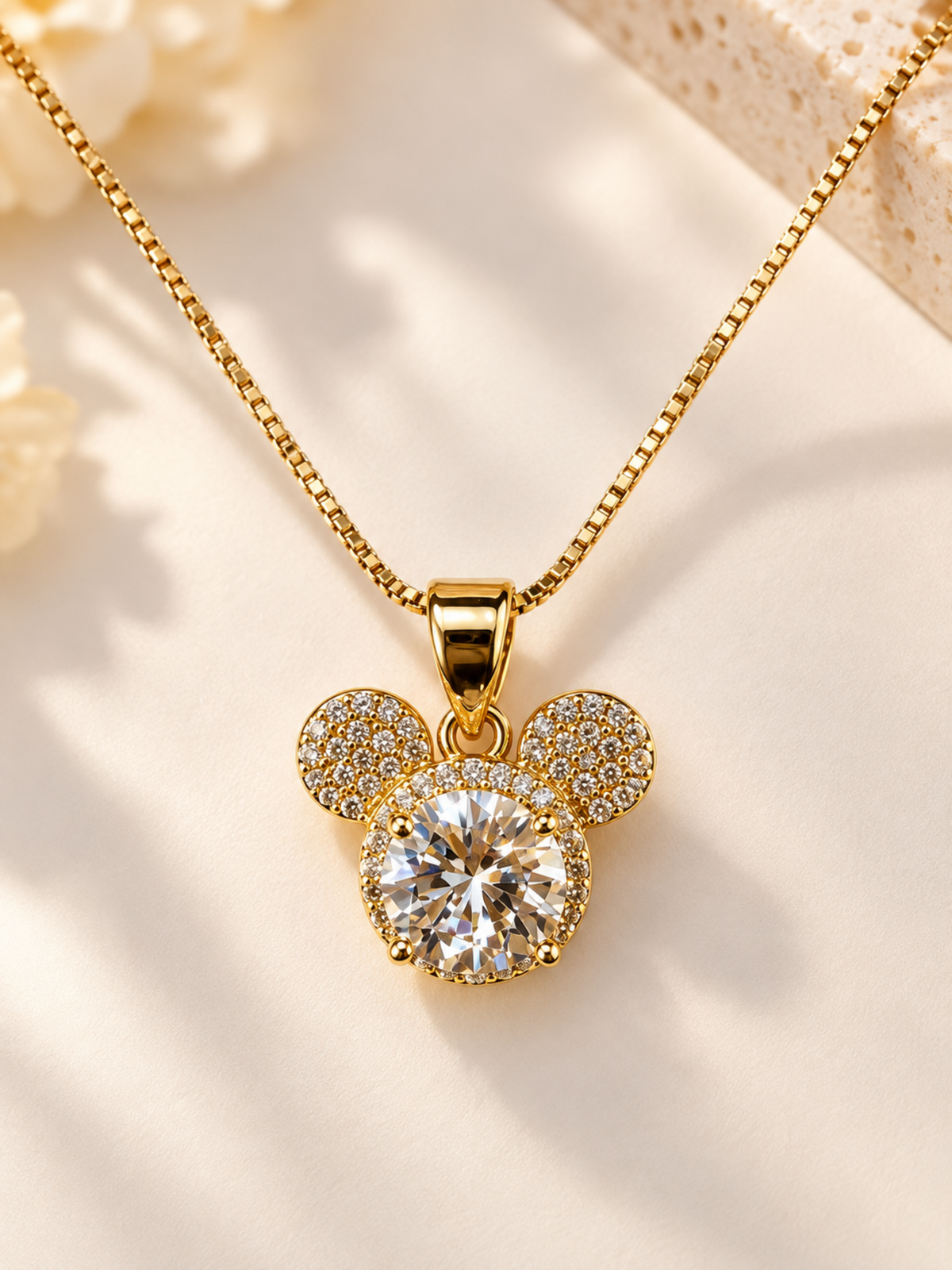 Gold-tone necklace with a Mickey Mouse-shaped pendant featuring a large clear round center stone and smaller clear stones around the ears, displayed on a warm cream surface.