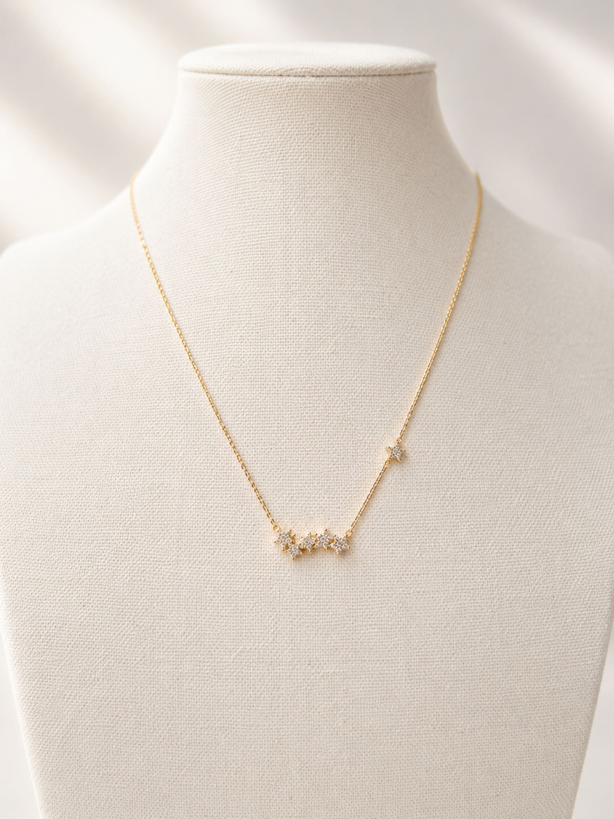 A delicate gold chain necklace with a small cluster of clear stones at the center and a tiny star charm on one side, displayed on a cream-colored fabric jewelry bust.