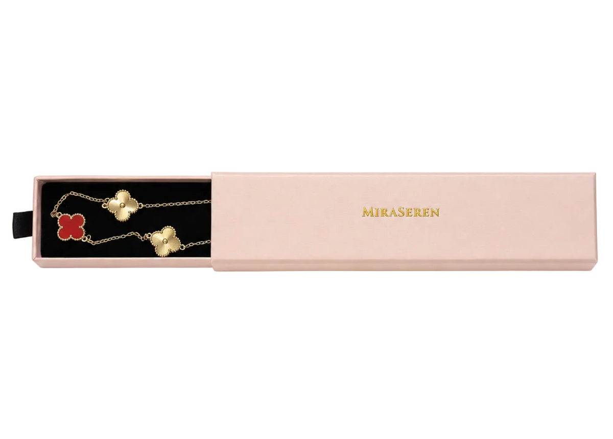 A pink rectangular jewelry gift box on a white background, with the left side slid open to reveal a black-lined compartment holding a gold-tone chain necklace with a red clover and white clover charm. The closed lid shows the brand name 'MIKASEREN' in gold.