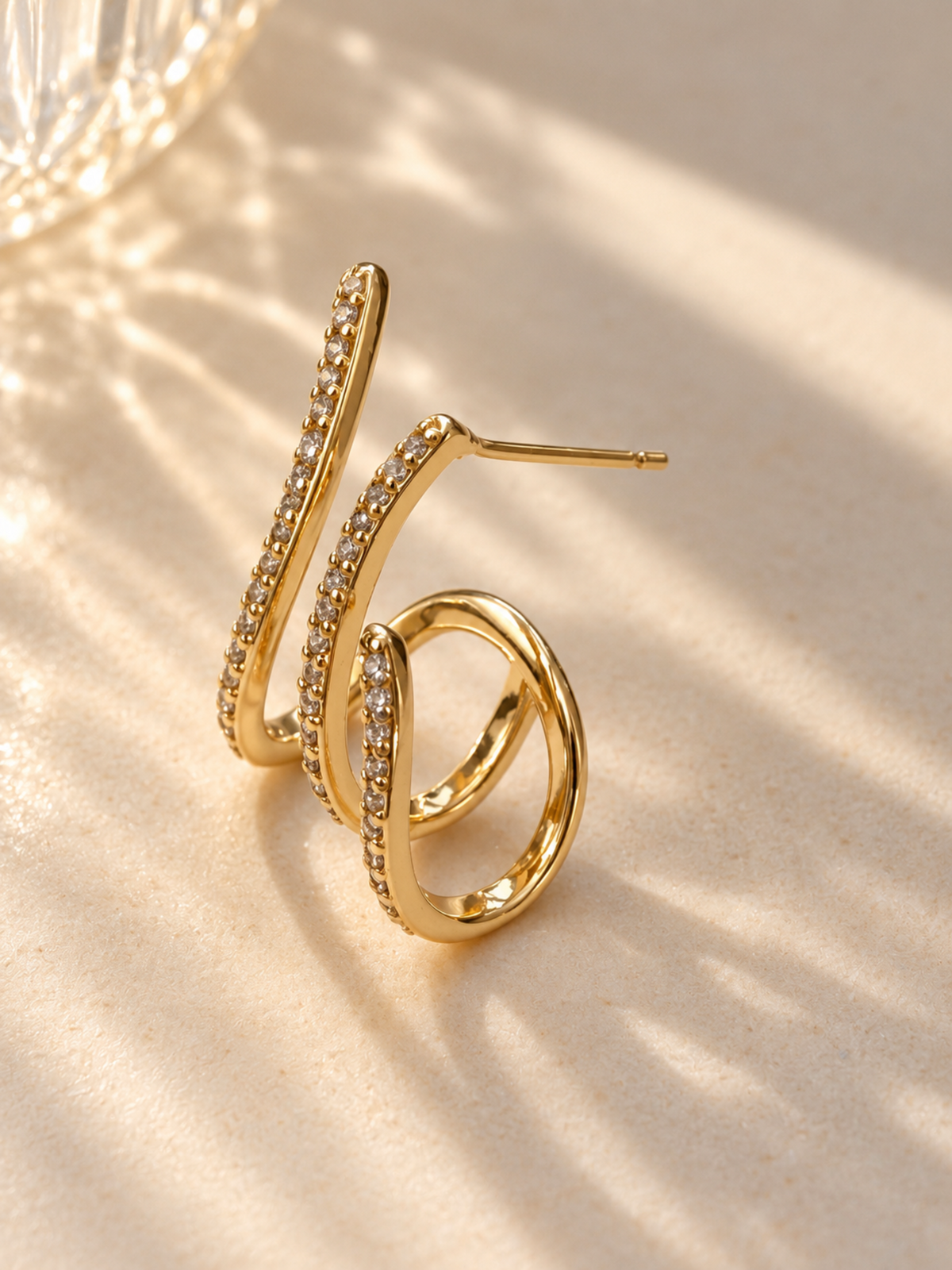 A pair of gold-plated spiral hoop earrings adorned with small clear pavé-set stones, displayed on a textured beige surface with soft, artistic shadows.