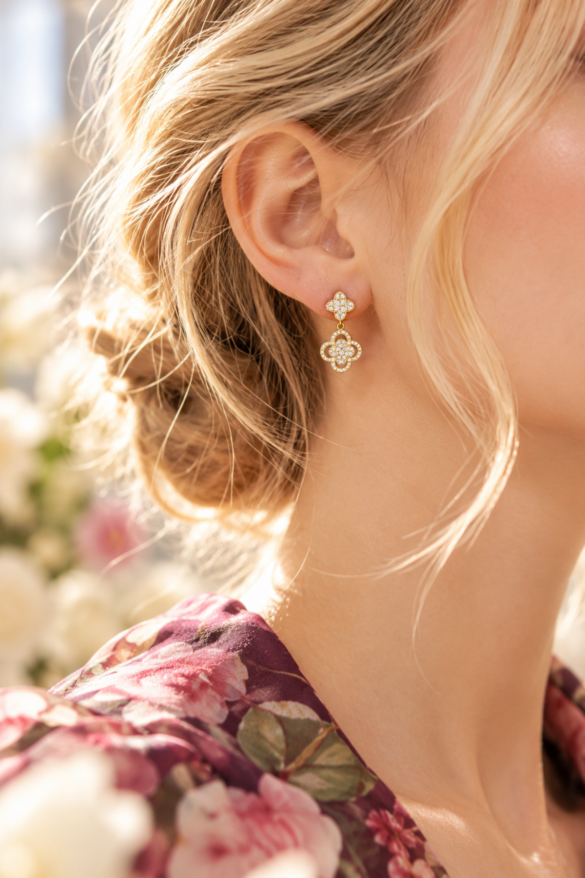 A close-up lifestyle shot of a woman wearing a delicate gold dangle earring with a four-leaf clover motif and small clear stones, set against a soft-focus background of white flowers.