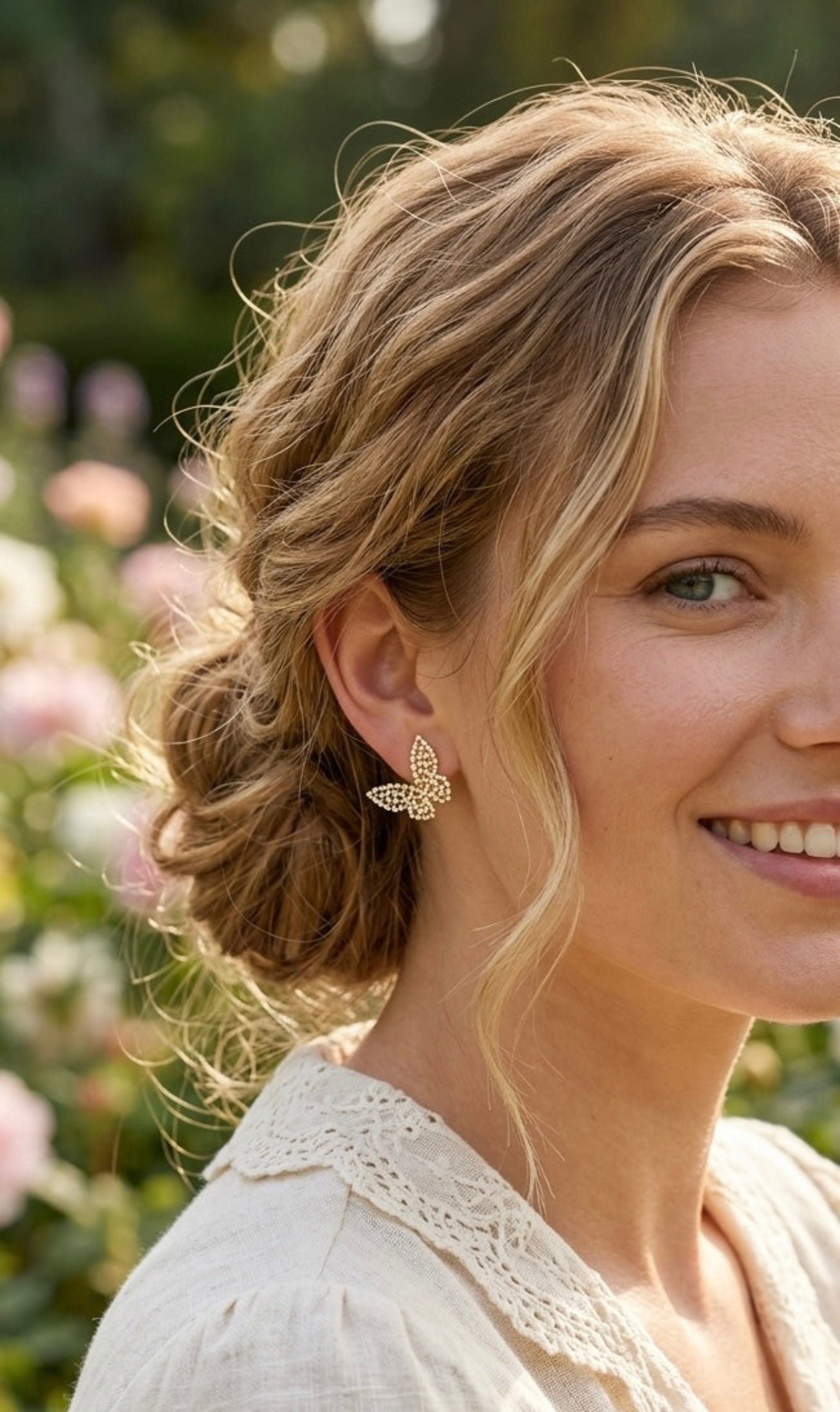 A close-up lifestyle shot of a woman with blonde hair wearing a delicate gold butterfly-shaped stud earring, set against a soft-focus garden background.