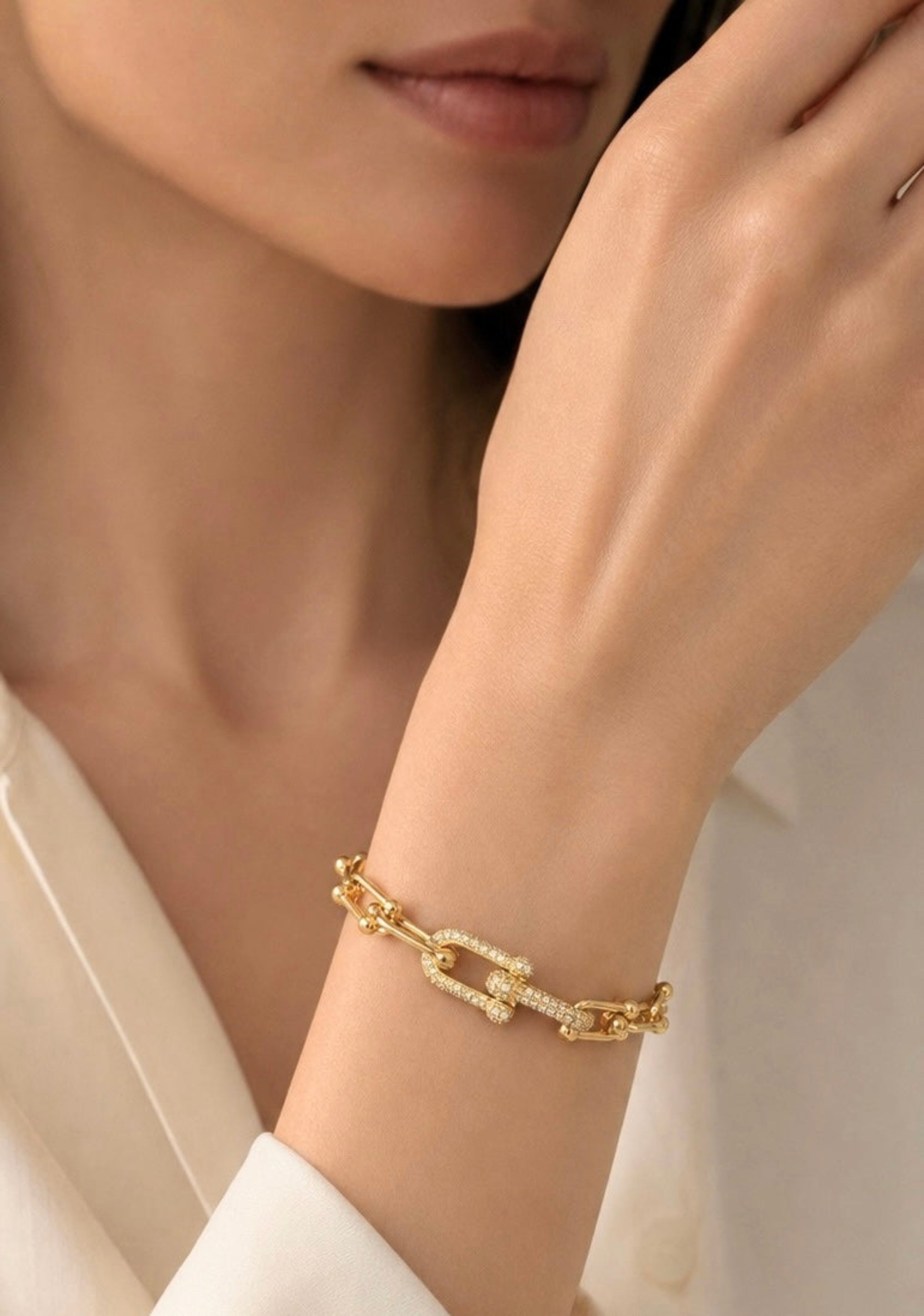 A close-up of a model's wrist wearing a gold chain link bracelet adorned with small clear stones, paired with a white collared shirt.