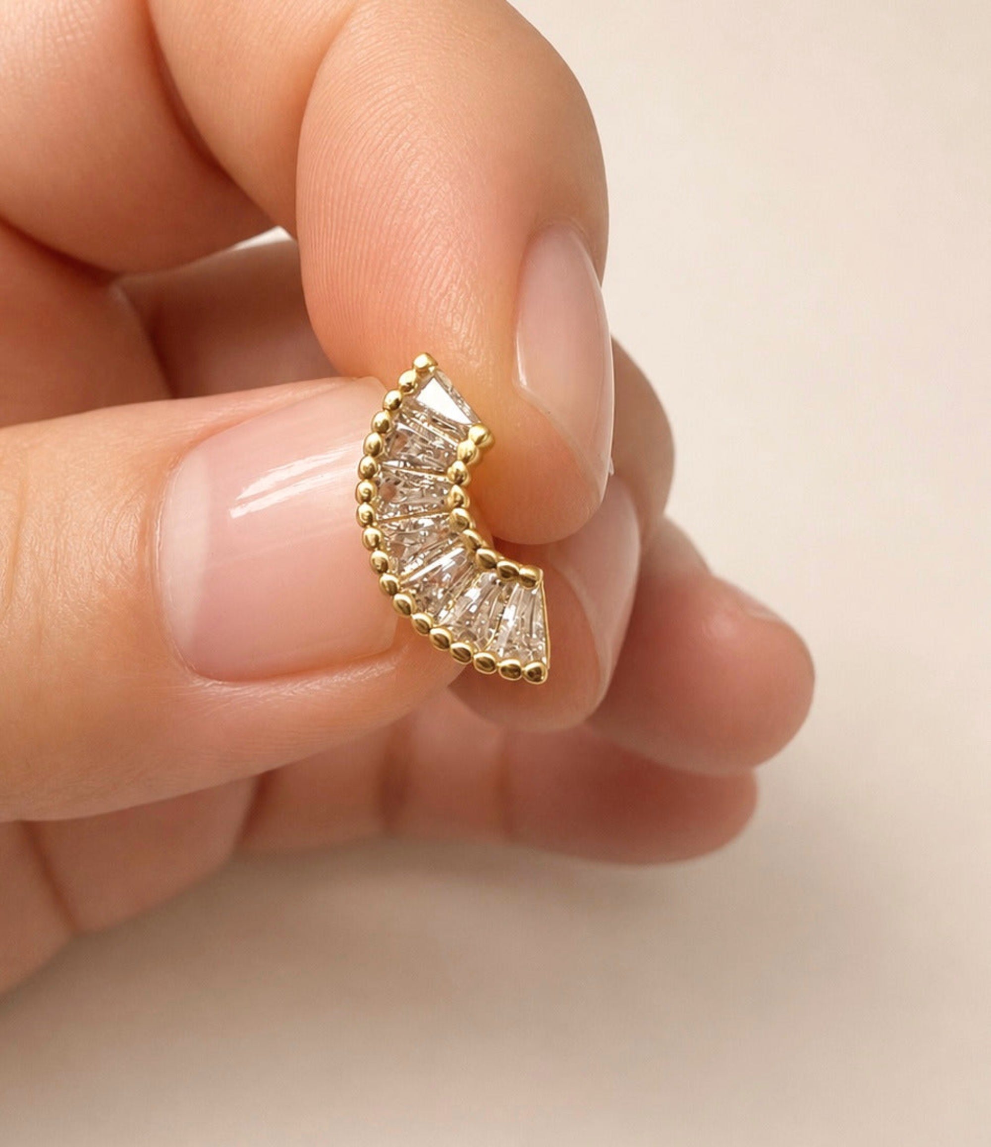 A close-up of a hand holding a small gold crescent-shaped stud earring set with clear baguette and round-cut stones against a soft beige background.