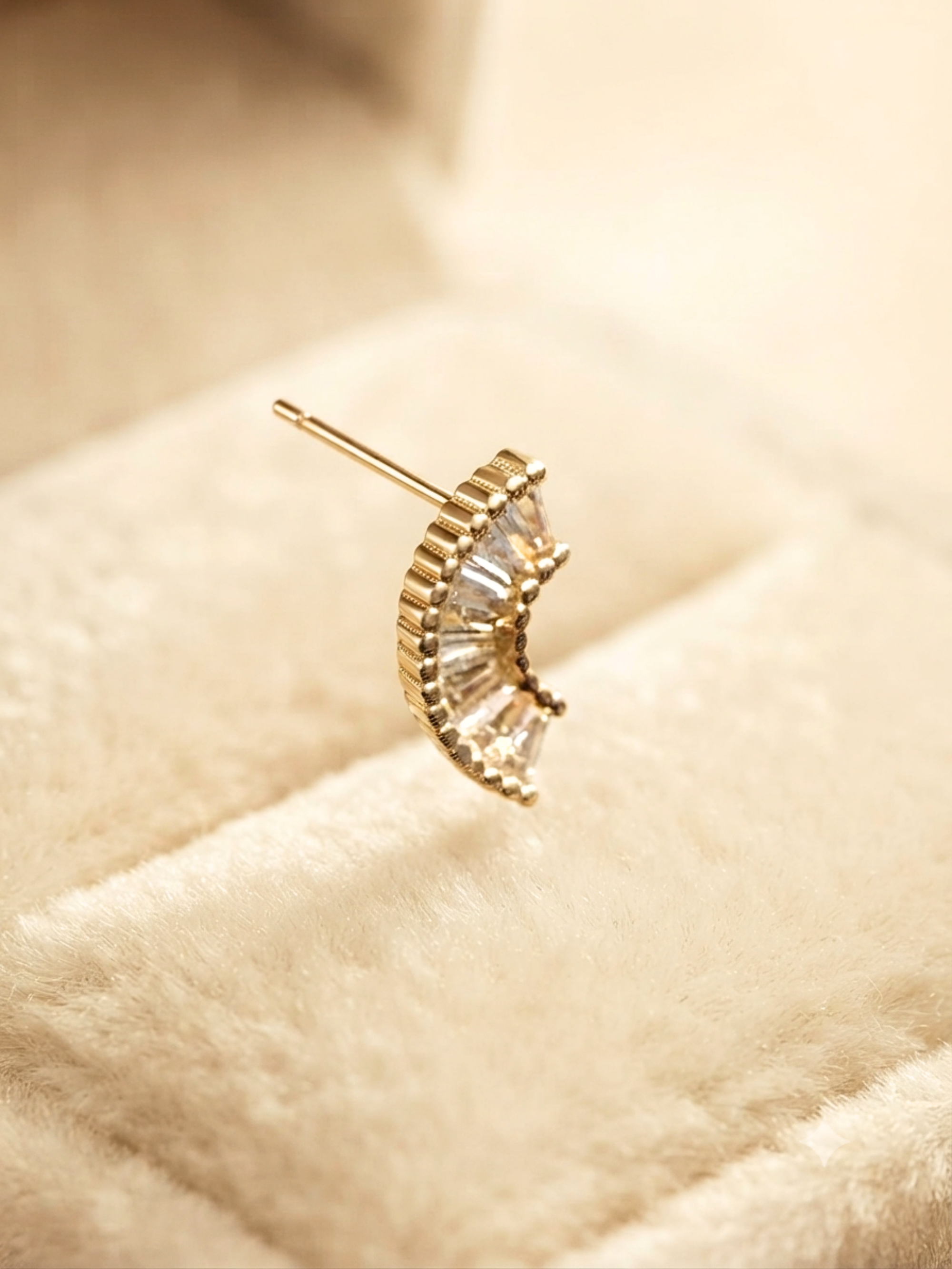 A gold crescent moon stud earring with clear baguette-cut crystals, resting on a soft beige velvet jewelry box.