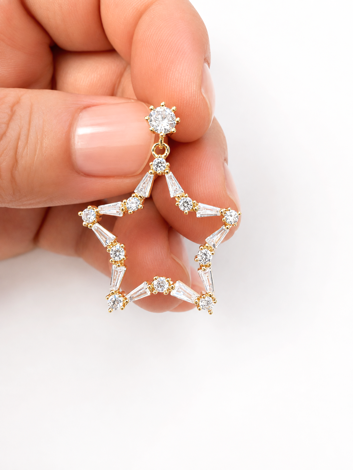 A hand with manicured nails holds a gold star-shaped dangle earring adorned with clear baguette and round-cut stones against a plain white background.