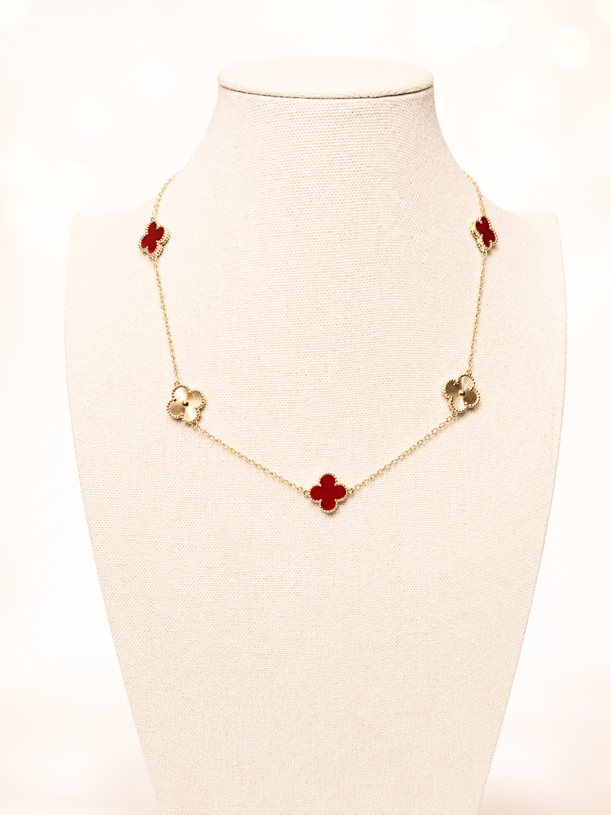 A gold-tone chain necklace with five red clover motifs, displayed on a cream-colored fabric jewelry bust against a bright, softly blurred background.