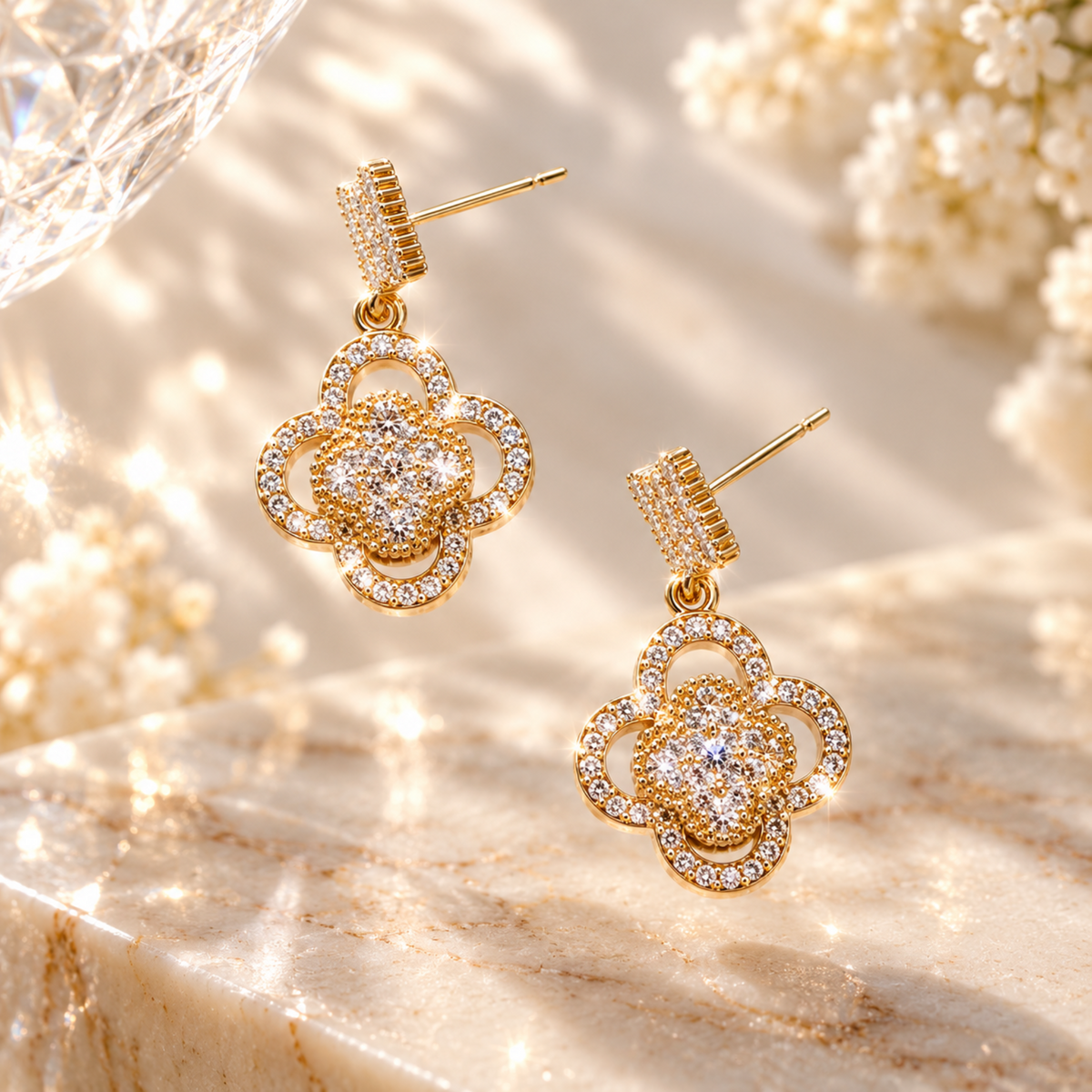 A pair of gold-toned dangle earrings with a floral clover design, embellished with clear crystals, displayed on a beige marble surface with soft-focus white flowers in the background.