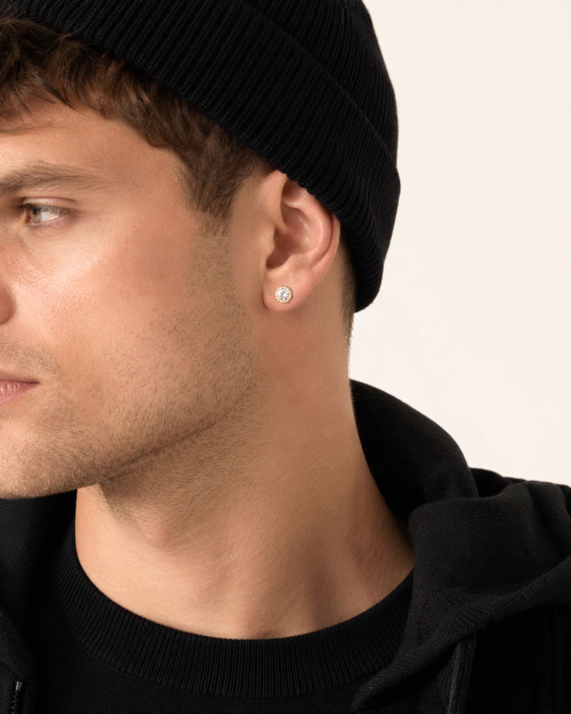 A close-up side profile of a man wearing a black beanie, black hoodie, and a small round silver stud earring with a clear gemstone on his earlobe, set against a plain off-white background.
