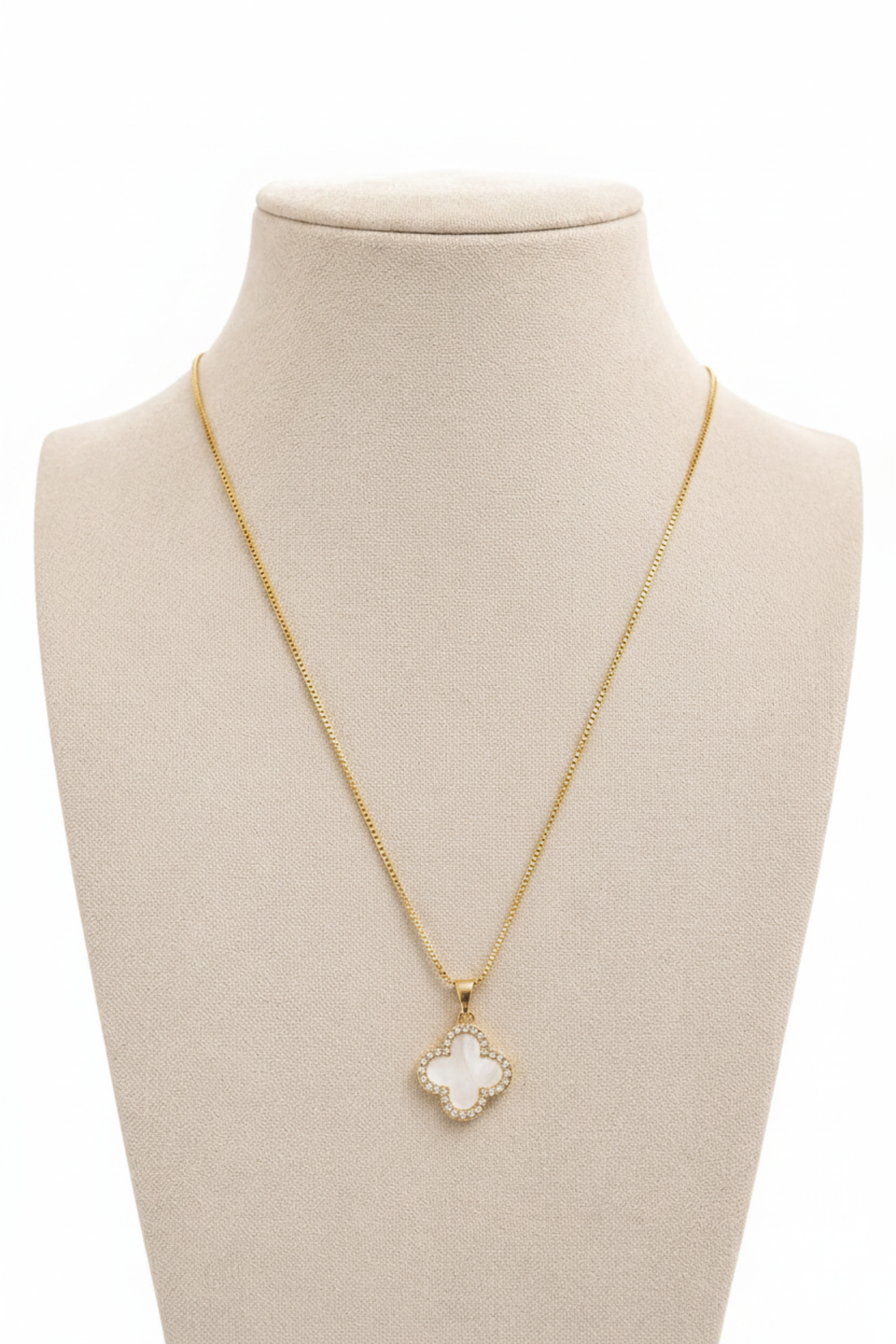 A delicate gold chain necklace with a four-leaf clover pendant, displayed on a beige fabric jewelry bust against a soft, light background.