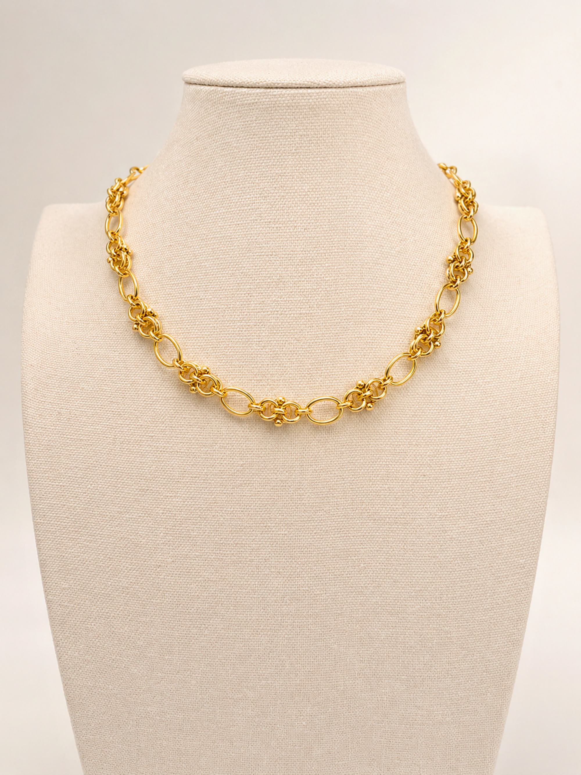 A gold chain necklace with alternating textured floral links and smooth oval links, displayed on a cream fabric jewelry bust against a light gray background.