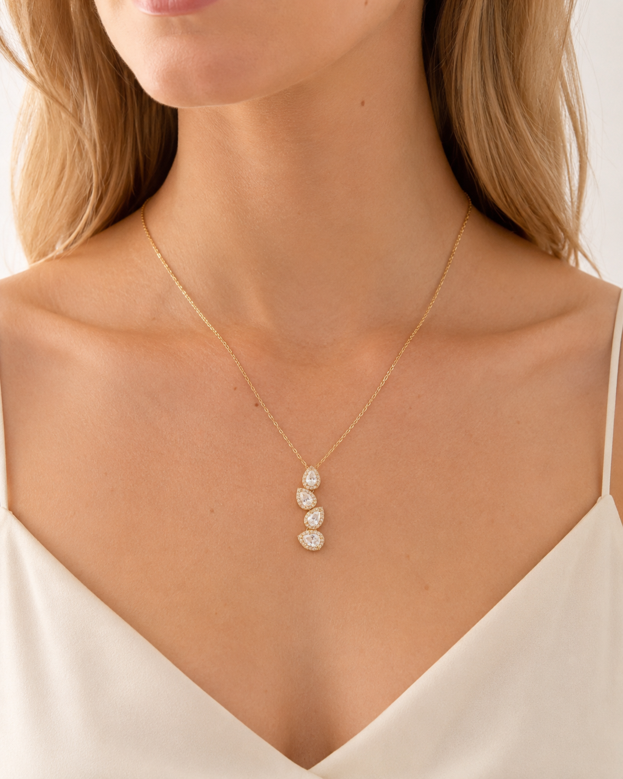 A close-up of a model wearing a delicate gold chain necklace with three small, clear teardrop-shaped stones set in gold, styled with a white camisole top.