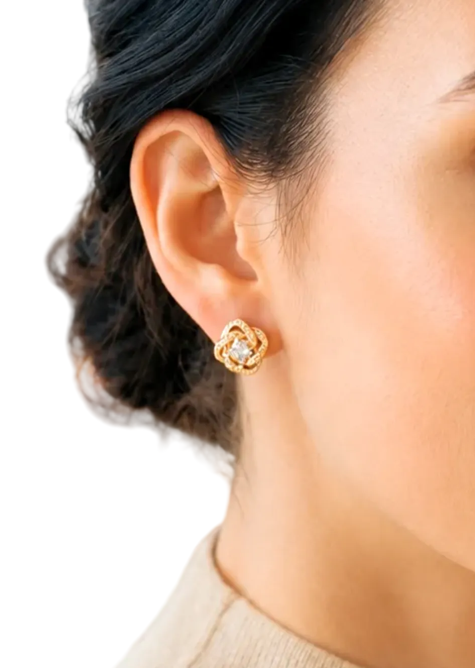 A close-up lifestyle shot of a woman with dark hair pulled back, wearing a small gold floral stud earring with clear stones. She is wearing a beige knit turtleneck sweater, with a softly blurred living room background featuring a brown leather chair.