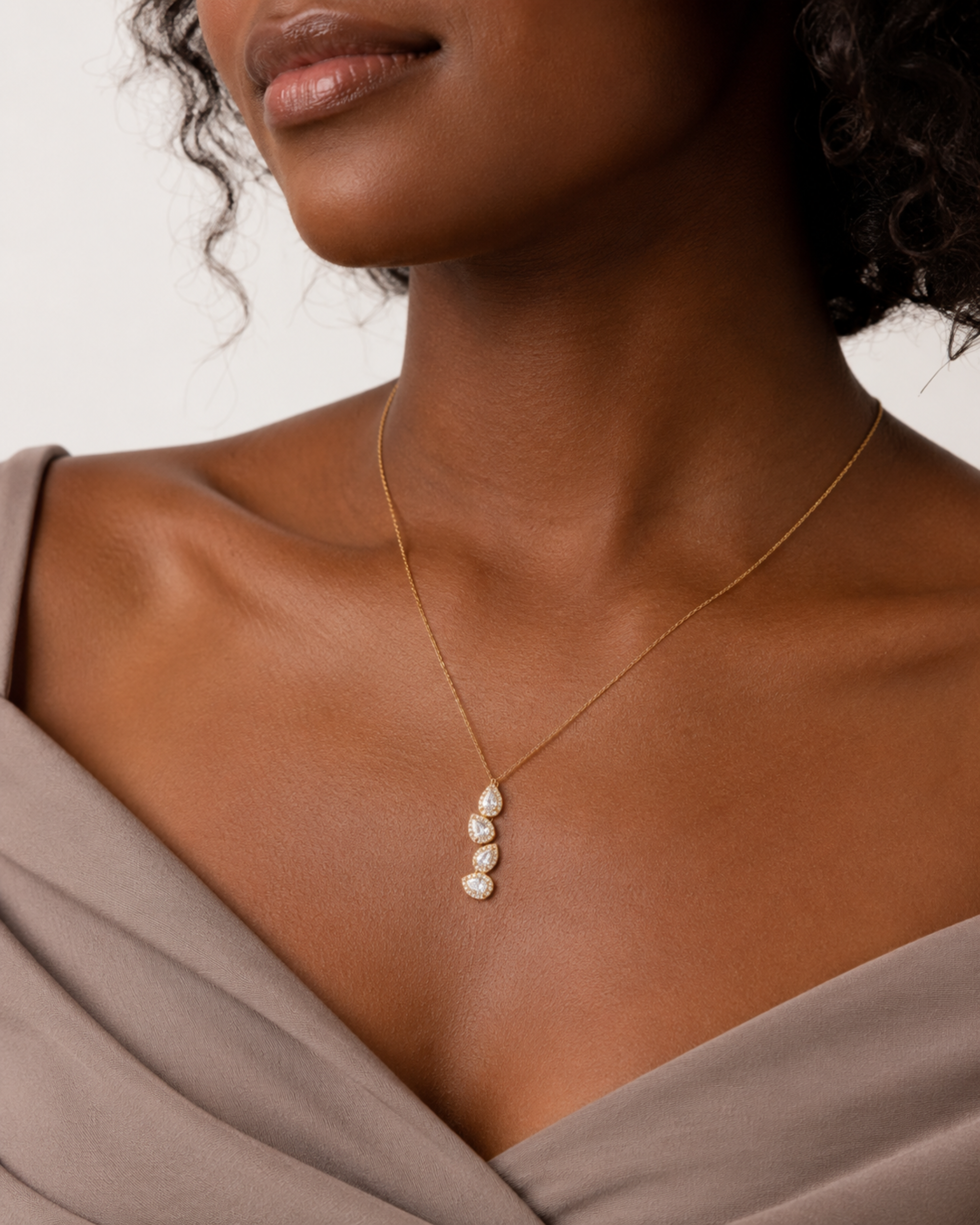 A close-up of a model wearing a delicate gold chain necklace with three small, pear-cut clear stones arranged vertically on a gold pendant, styled with a taupe off-the-shoulder top.