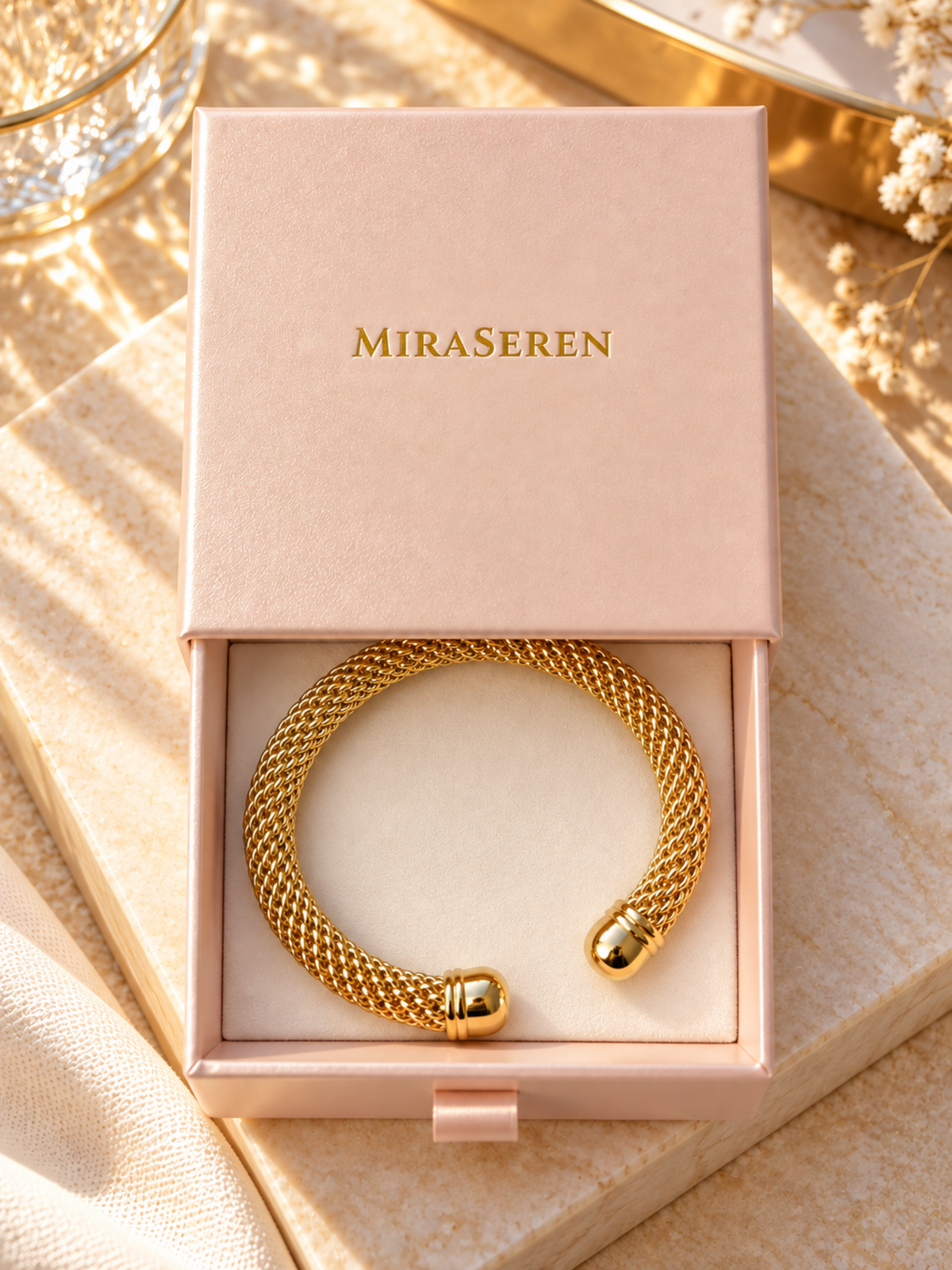 A gold-tone twisted cable cuff bracelet by MiraSeren, presented inside a light pink jewelry box on a warm beige textured surface.