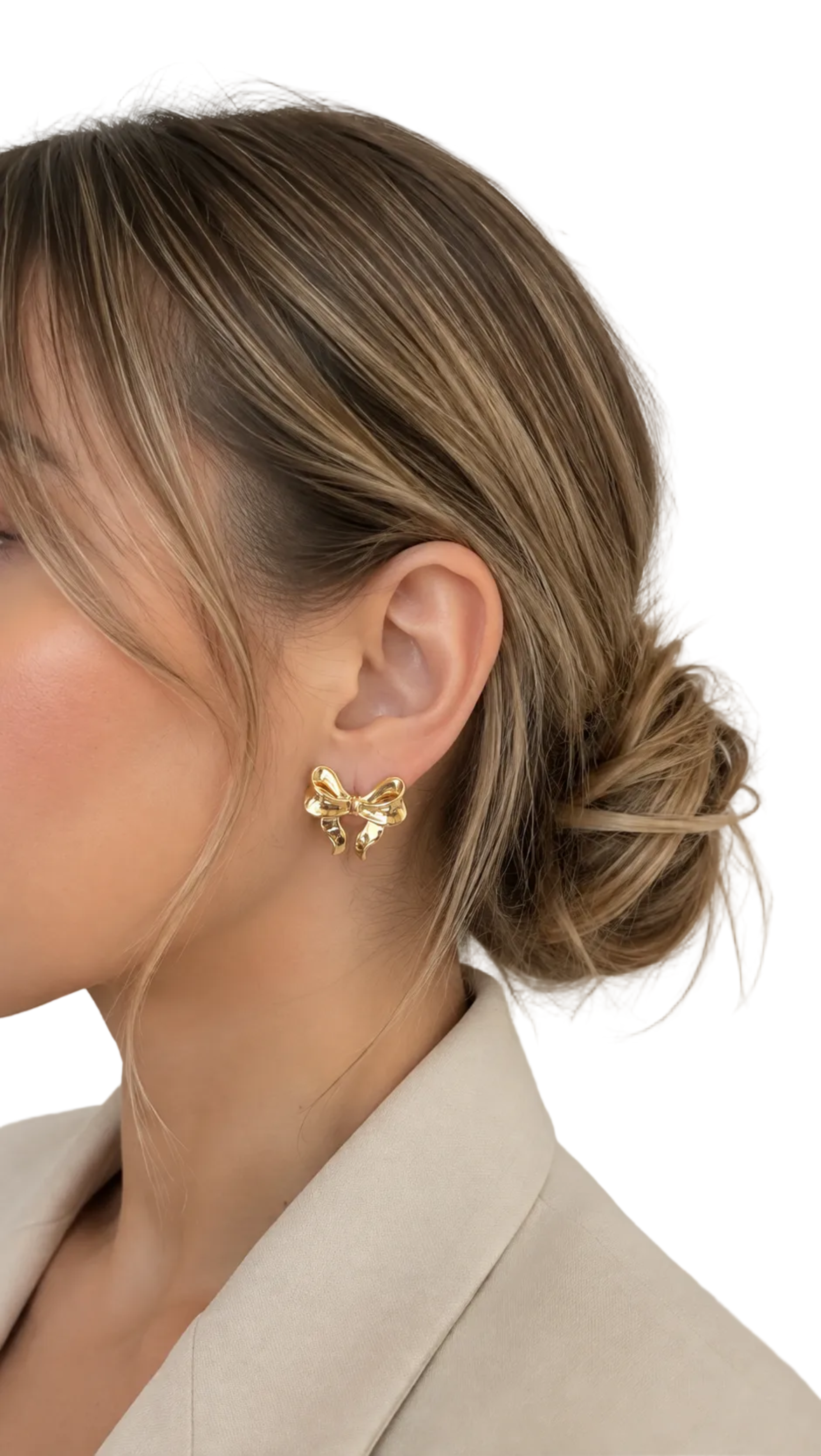 A close-up side profile of a woman with blonde-highlighted hair in a low bun, wearing a small gold bow-shaped stud earring and a light beige blazer against a plain light gray background.