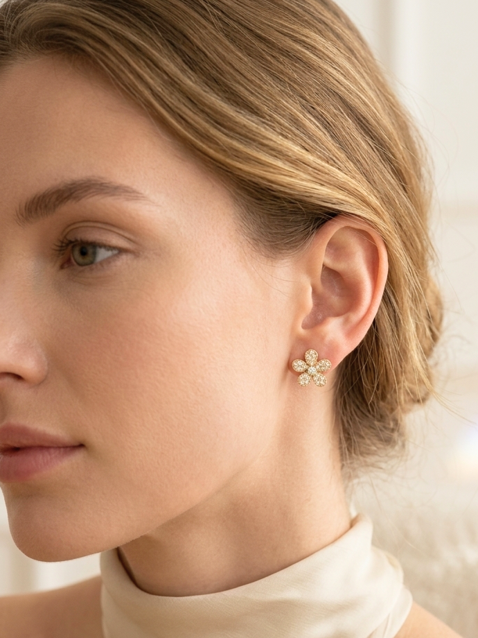 A close-up side profile of a woman with blonde hair wearing a small flower-shaped stud earring with clear stones in a gold-tone setting, paired with a cream satin high-neck top against a white background.