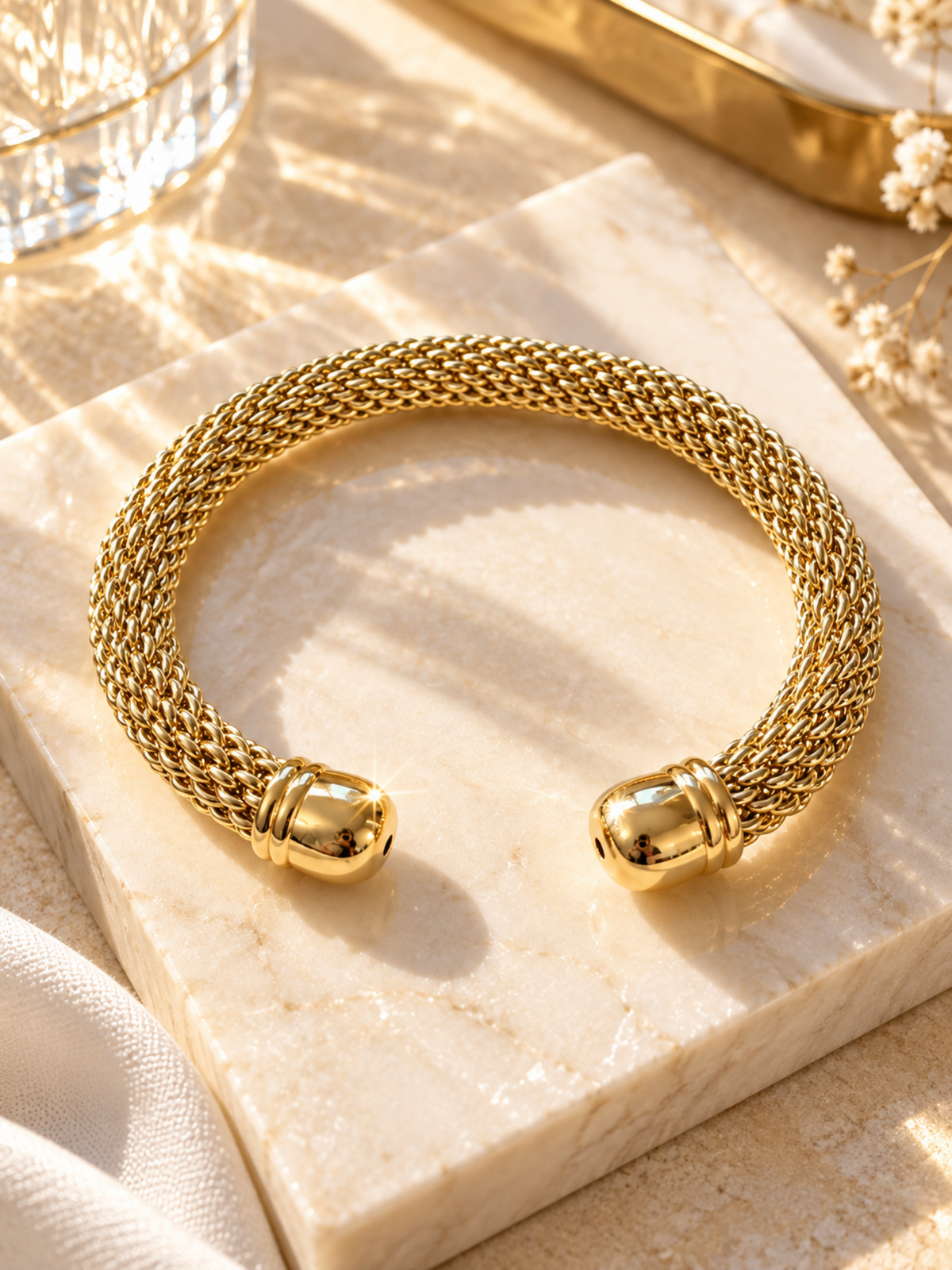 A gold-toned braided cuff bracelet displayed on a light marble block with warm sunlight and soft decorative props around it.