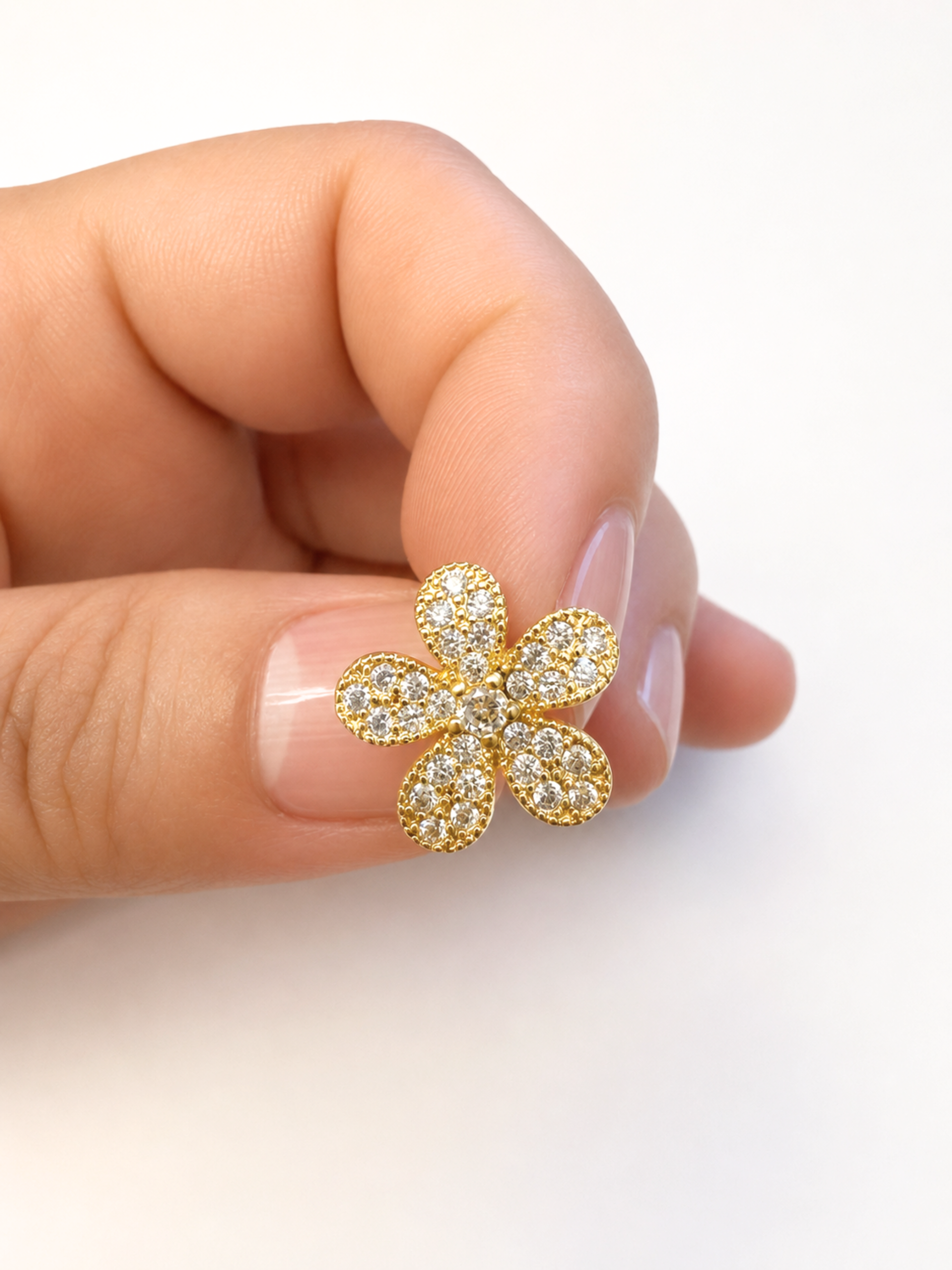 A gold flower-shaped stud earring paved with small clear stones, held between a person's thumb and index finger against a clean white background.