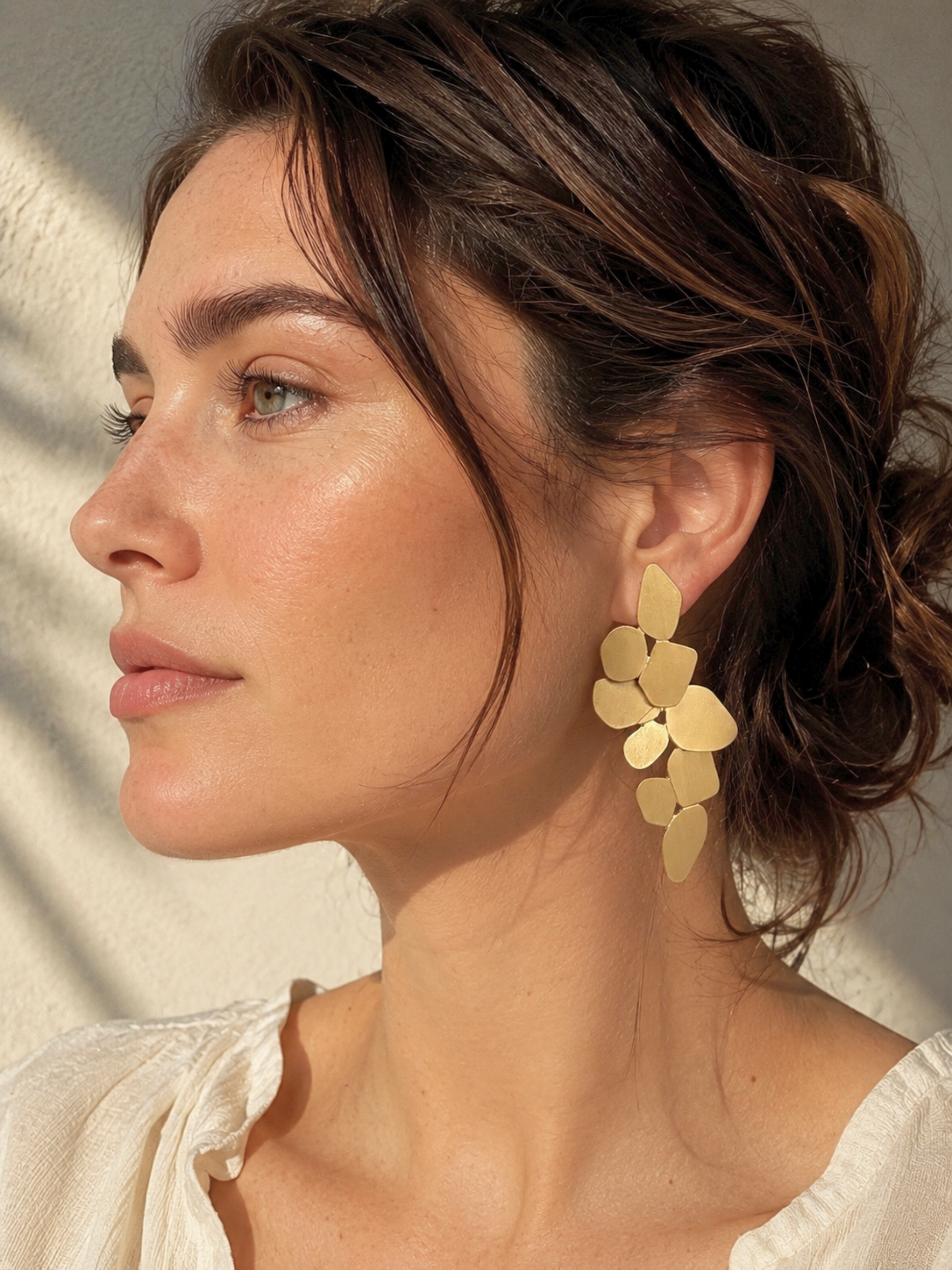 A woman in profile wearing a large gold-toned flower-shaped dangle earring, shown in warm natural light against a light neutral background.