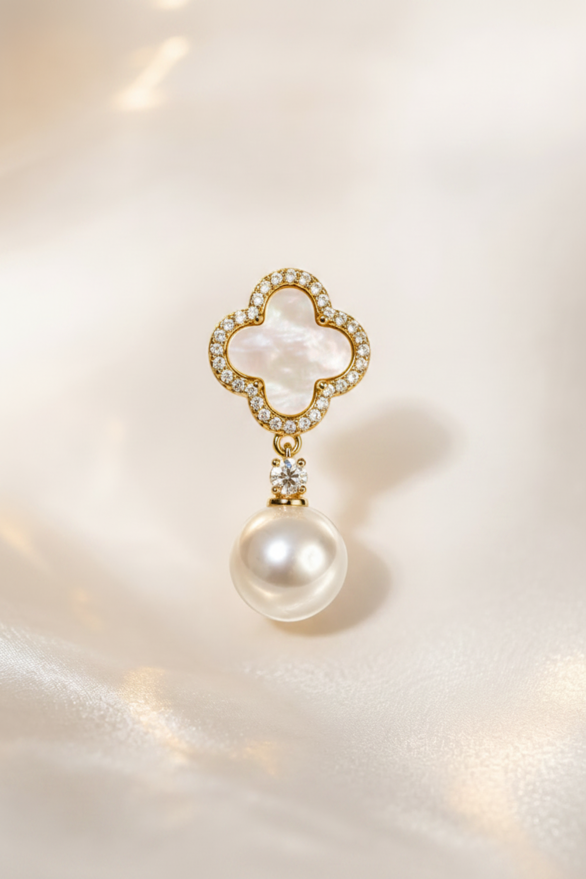 A single gold-toned drop earring featuring a four-petal clover motif with a white iridescent shell center, surrounded by small clear stones, with a dangling white pearl below, set against a soft cream satin background.