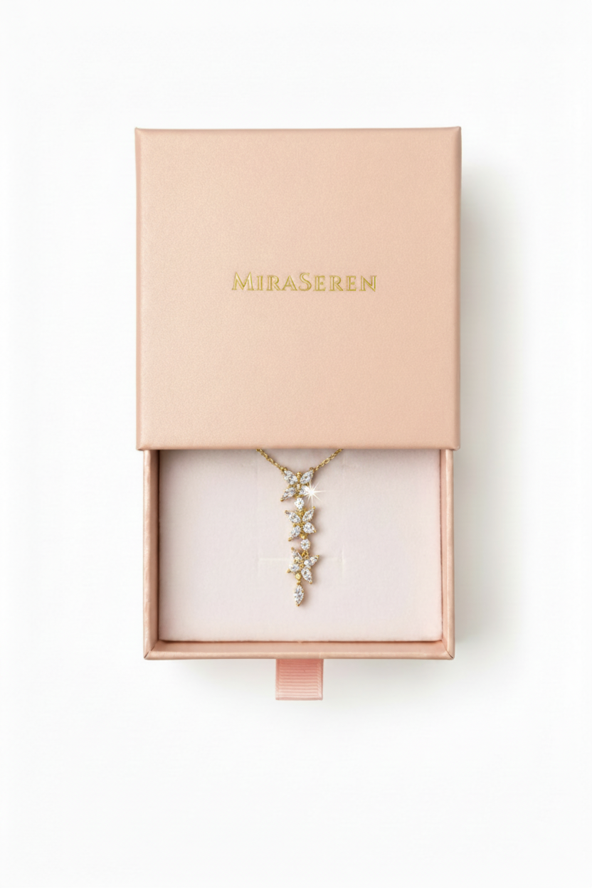 A gold-toned Miraseren necklace with a delicate floral drop pendant, presented inside an open blush pink Miraseren jewelry box on a clean white background.