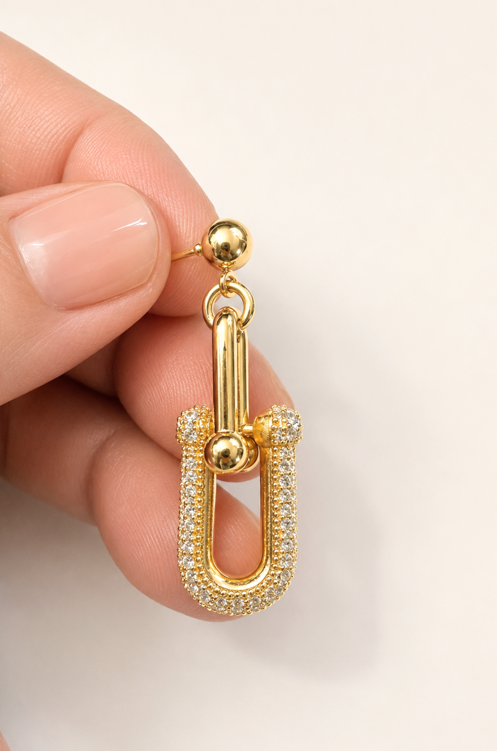 A gold U-shaped drop earring with pavé-set clear stones and a polished ball detail, held between fingers against a clean off-white background.